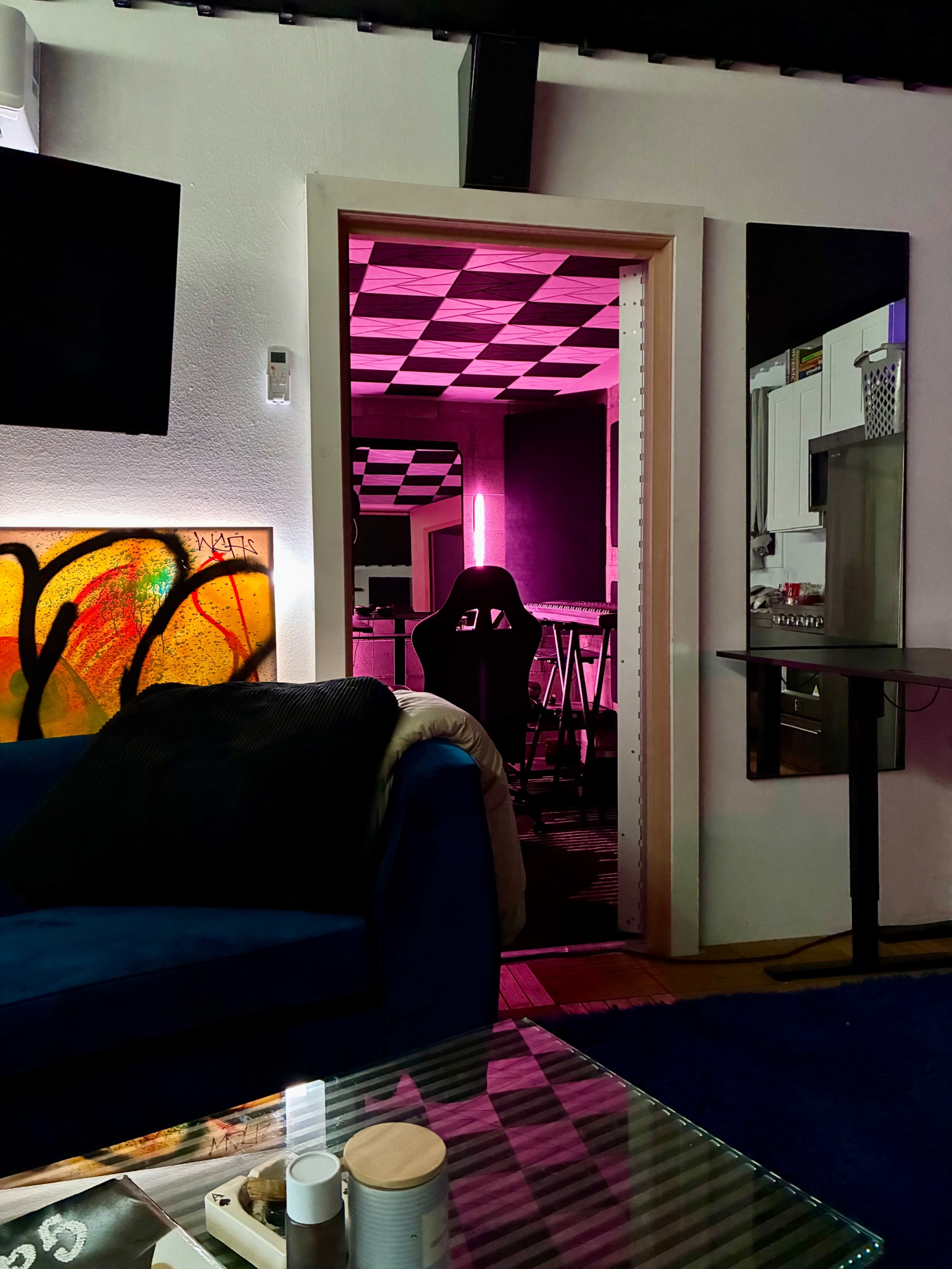 A brightly lit room with pink lighting is visible through an open door, contrasting with a dark, cozy living area.