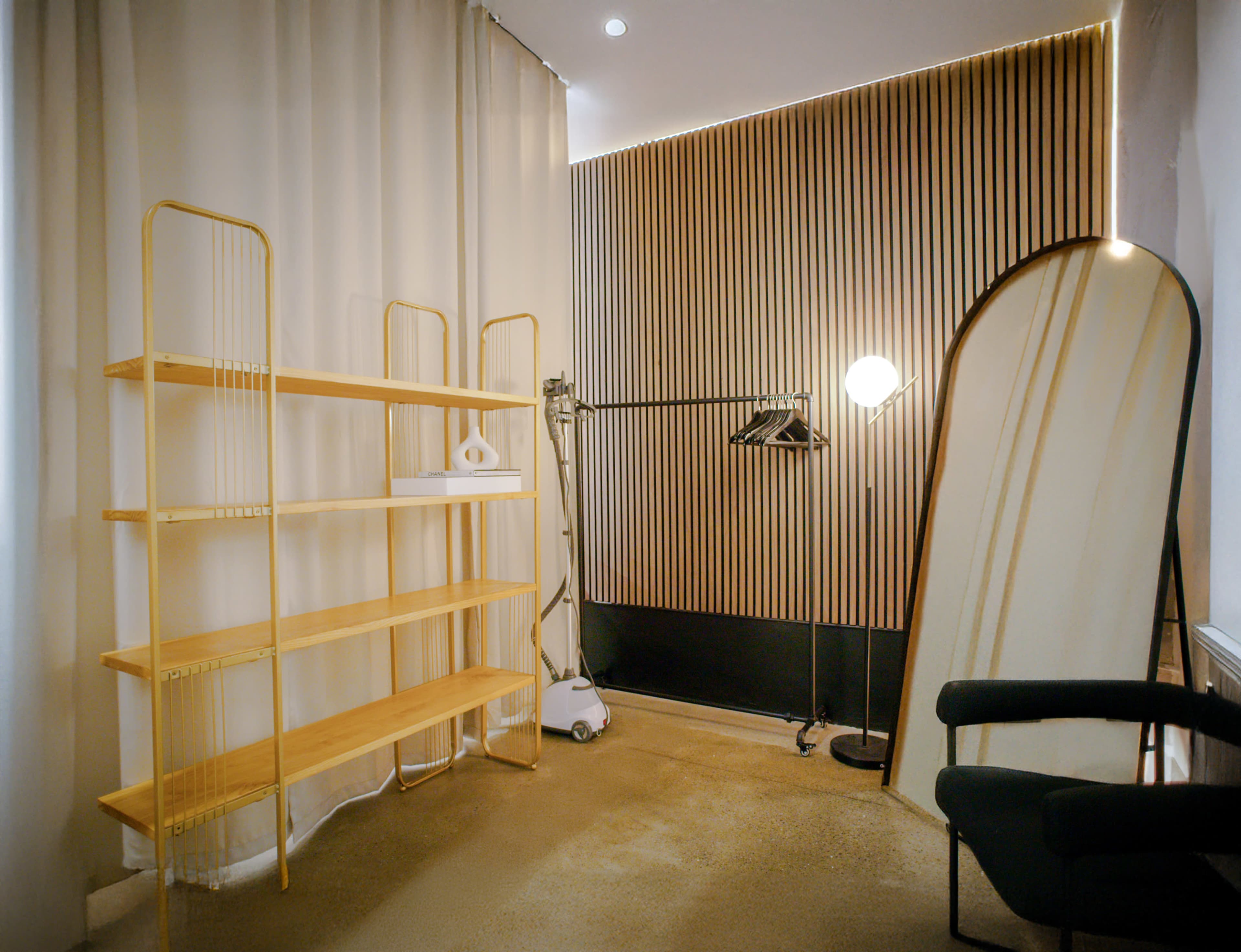 The image shows a minimalist interior featuring a wooden shelving unit, a clothing rack, a standing mirror, and a modern floor lamp against a wall with vertical wooden panels.