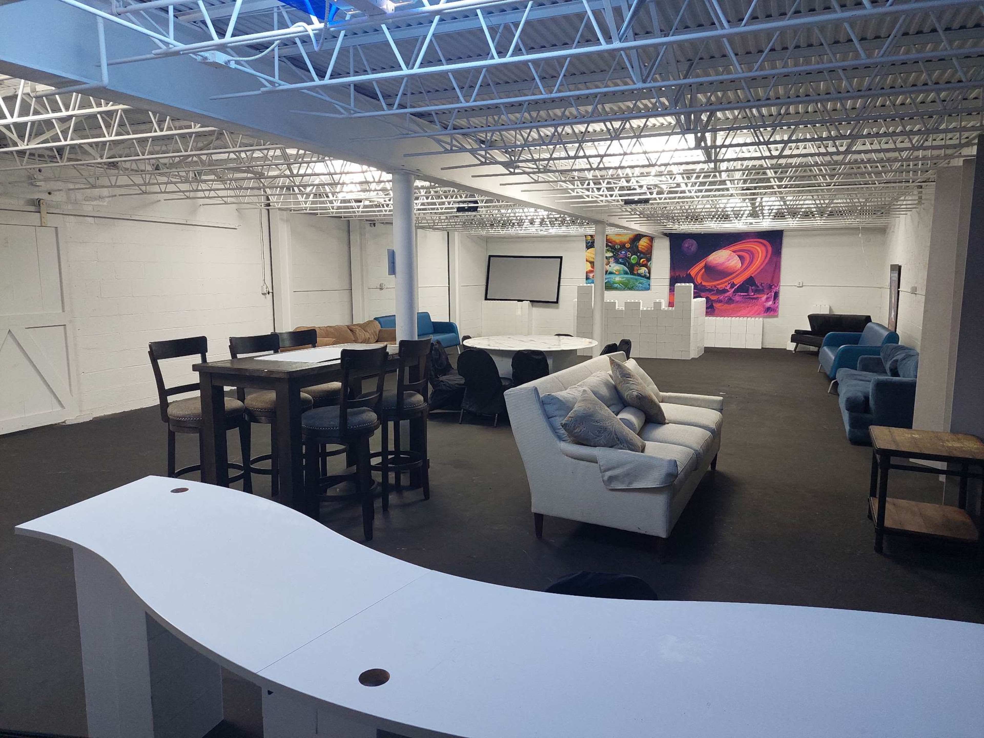 A spacious, well-lit communal area with various seating arrangements, including tables and couches, and a large screen at one end.