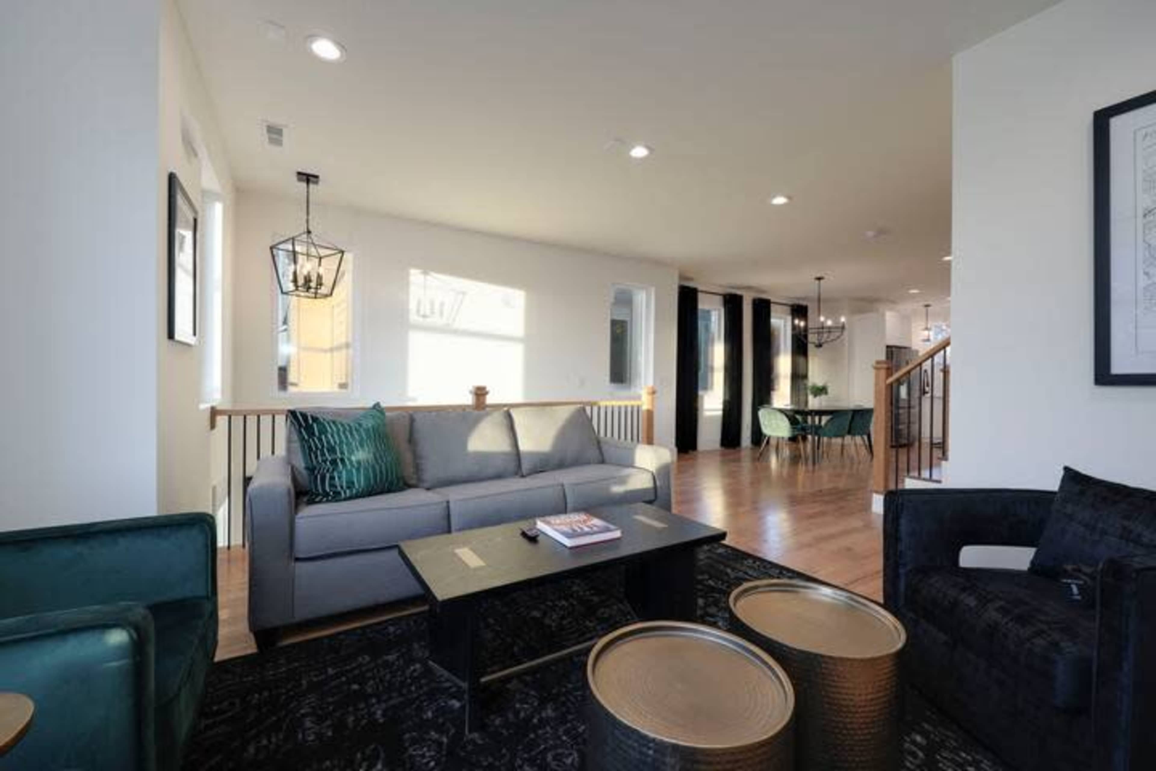 A modern living space with a gray sofa, dark decor, a wooden dining area, and large windows allowing natural light.