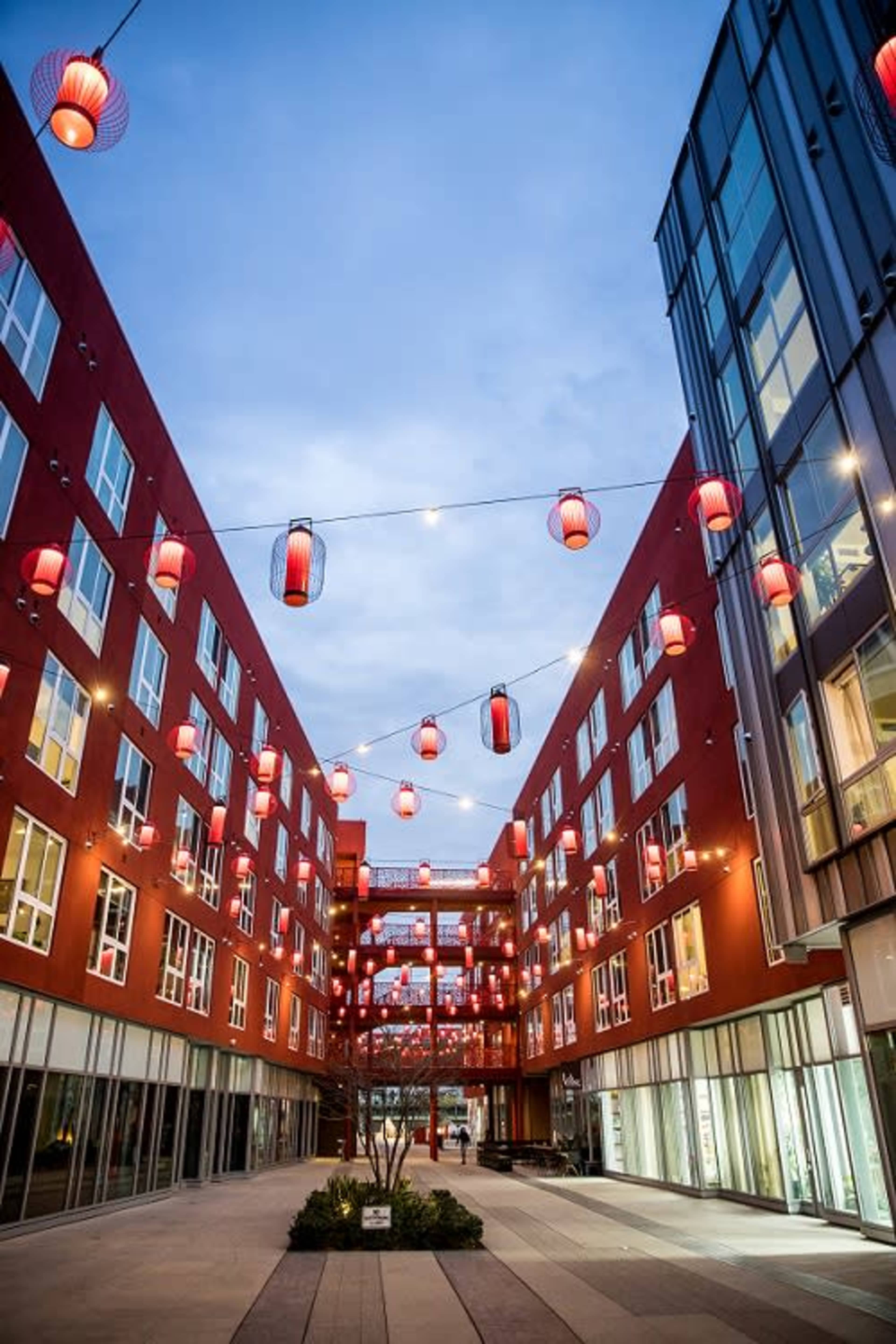 A narrow courtyard with red buildings and hanging lanterns is illuminated against a twilight sky.
