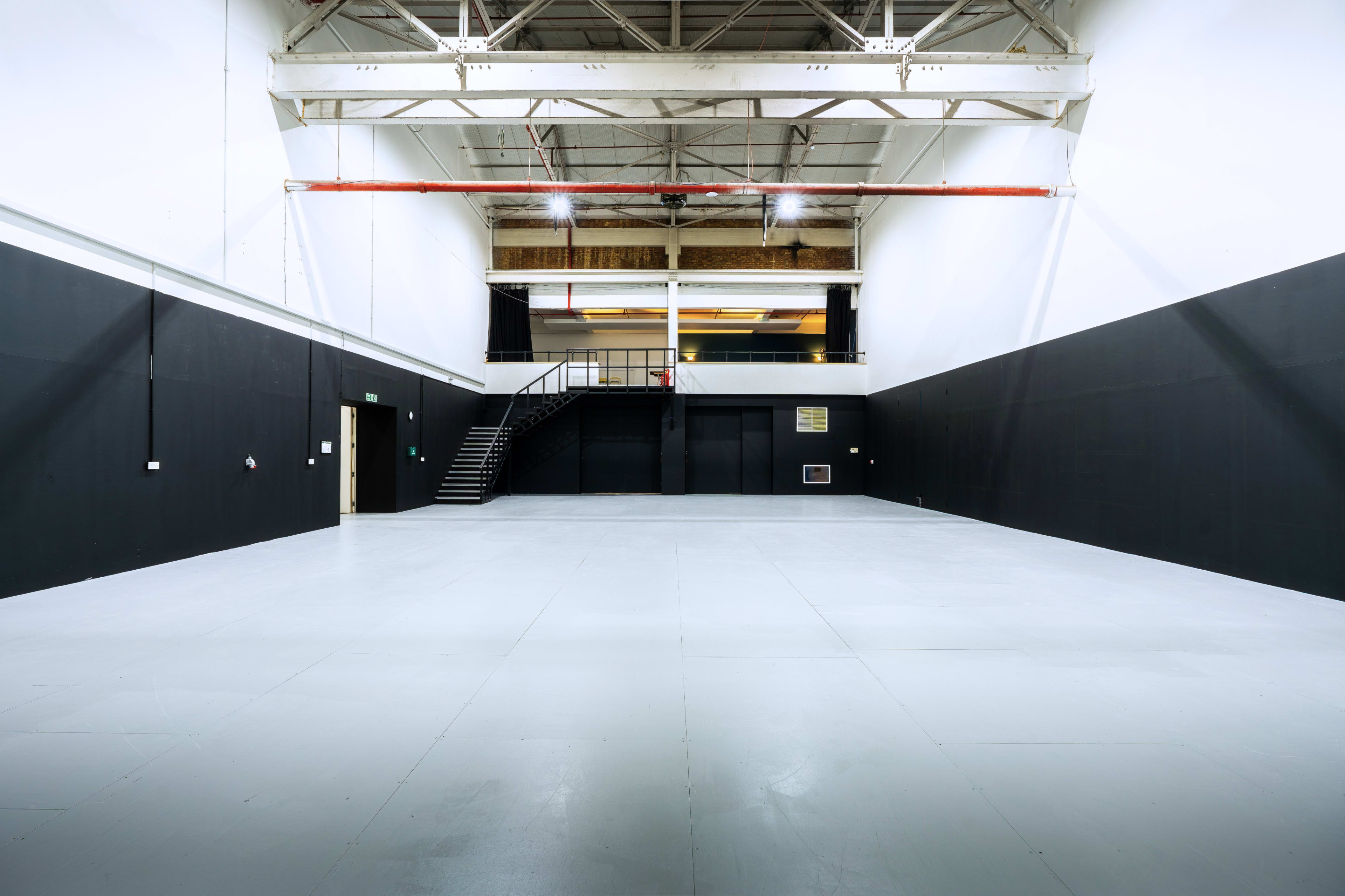 The image shows a large, empty industrial space with black walls and a white floor, featuring a staircase in the background.