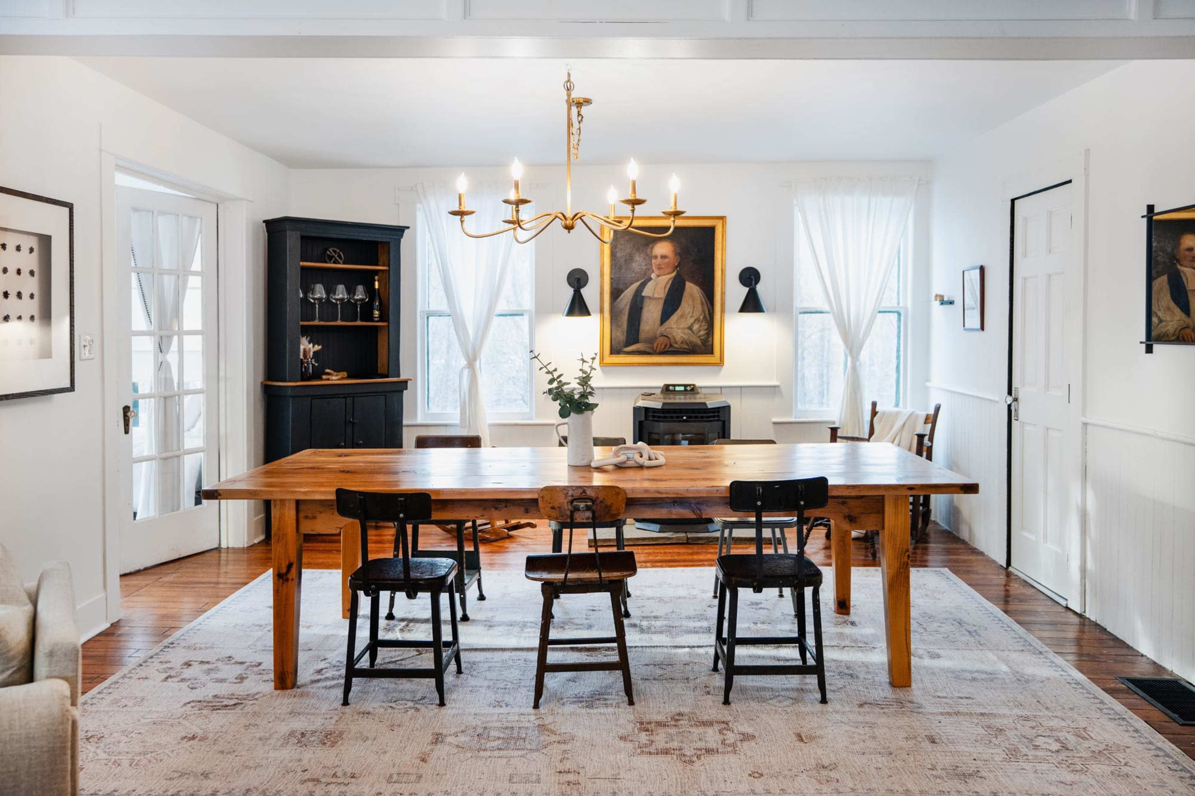 A dining room with a large wooden table surrounded by four chairs, framed by white walls and a portrait hanging above a black cabinet.
