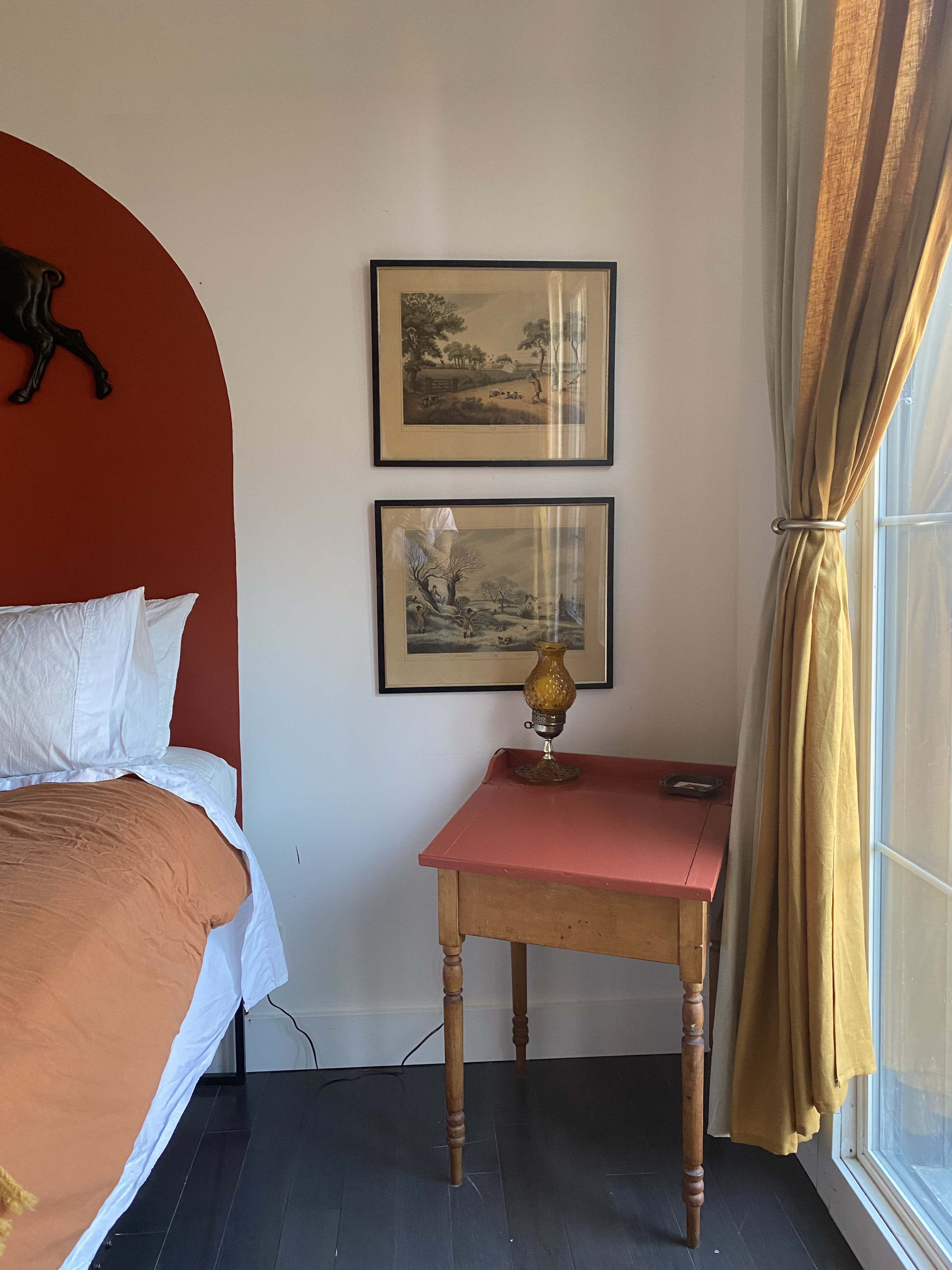 A bedroom corner featuring a bed with white linens, two framed pictures on the wall, a small table with a lamp, and a window with curtains.