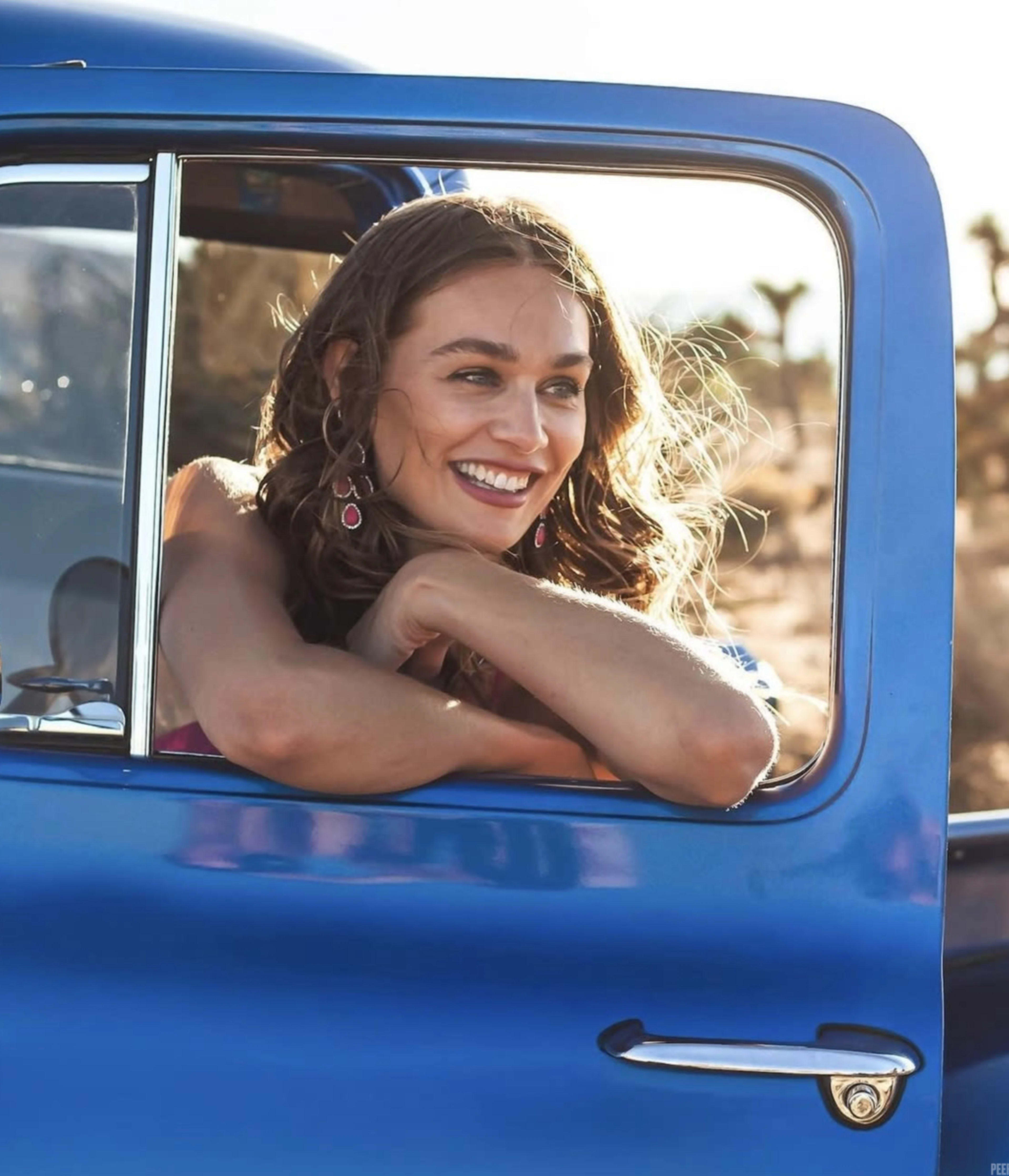 A woman with long hair leans out of the window of a blue vintage truck in a desert landscape.