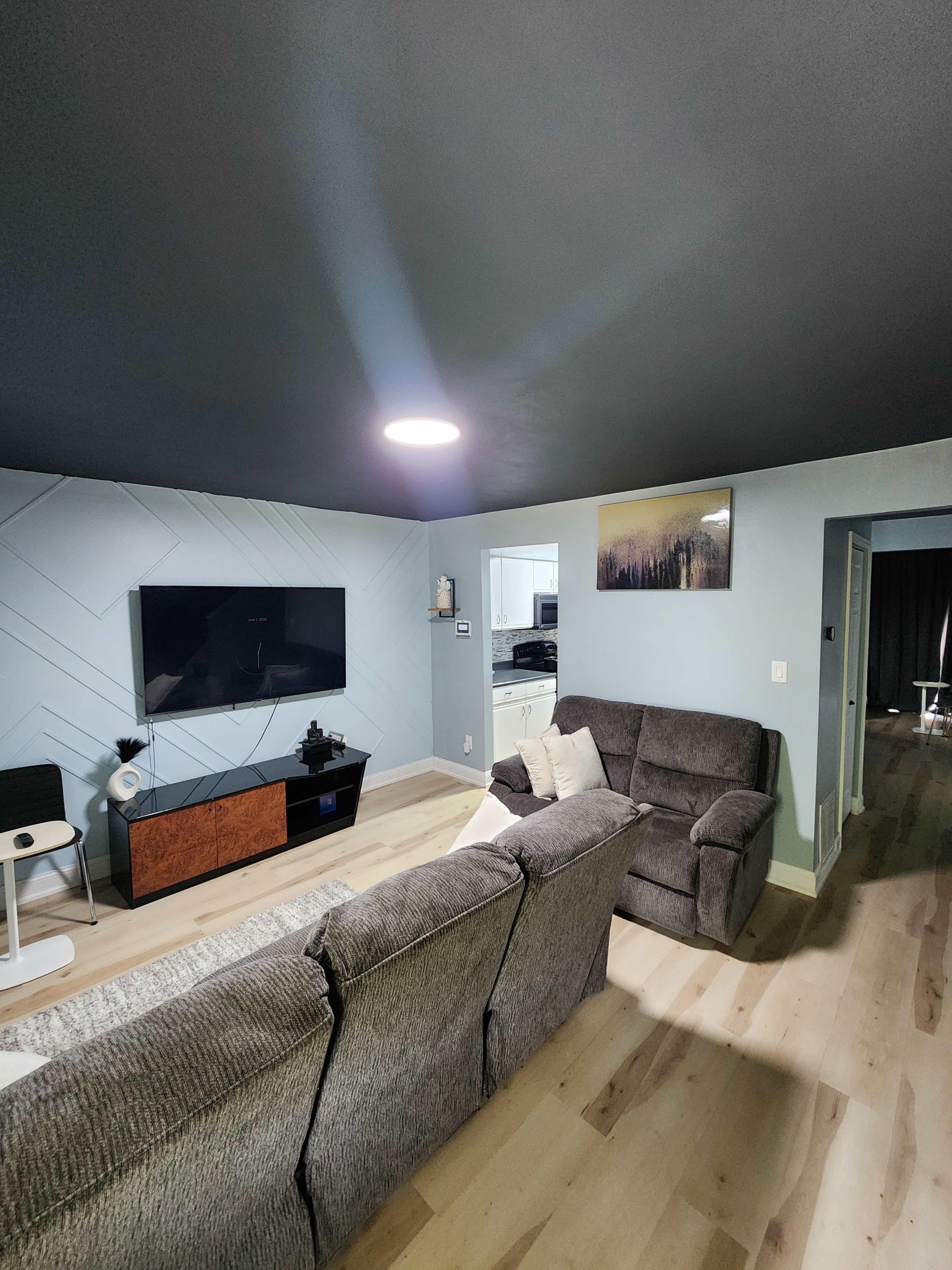 The image shows a living room with two sofas facing a wall-mounted television, light-colored walls, and a painted ceiling.