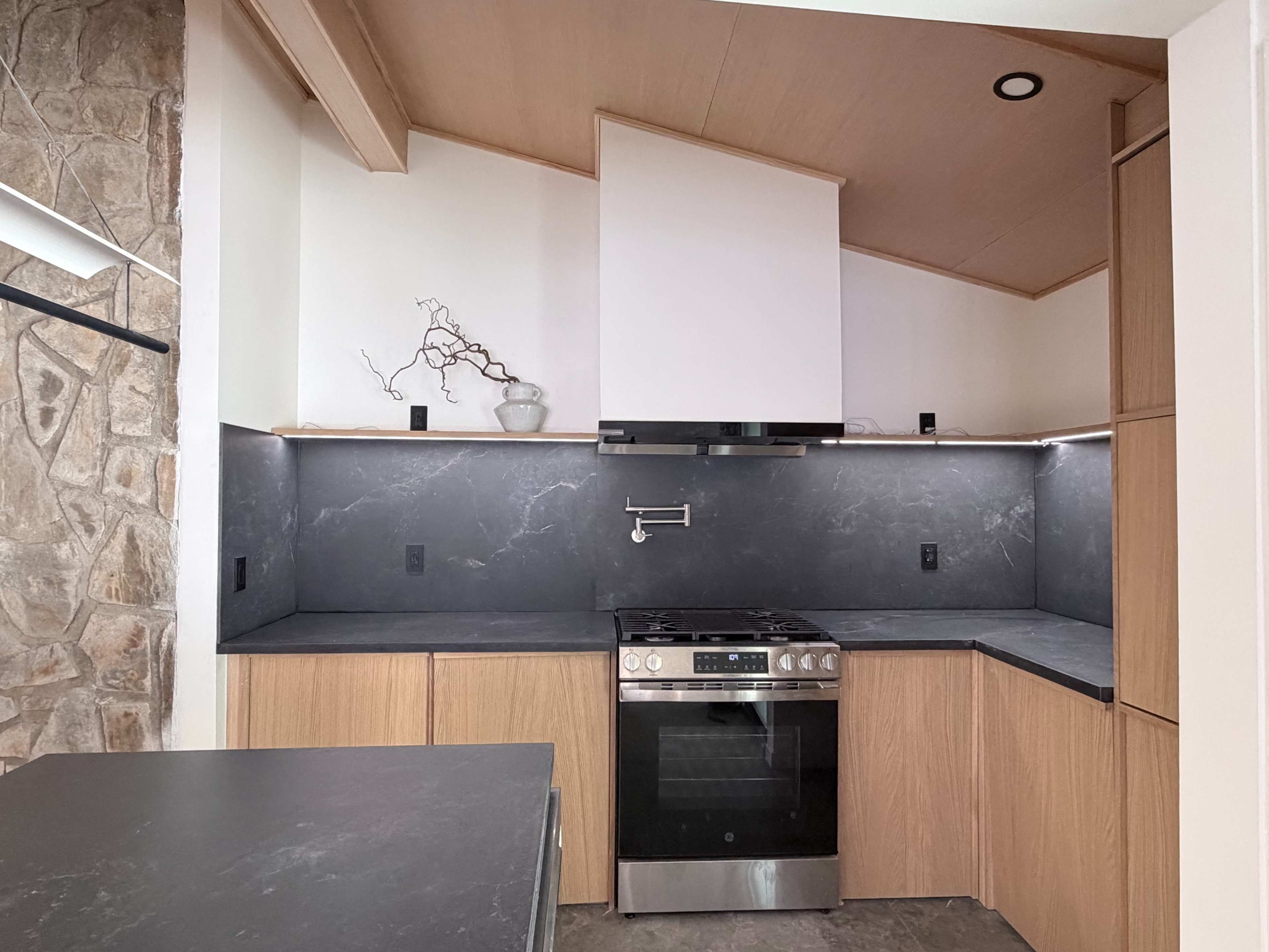 The image shows a modern kitchen featuring wooden cabinets, a stone backsplash, a stainless steel stove, and dark countertops.