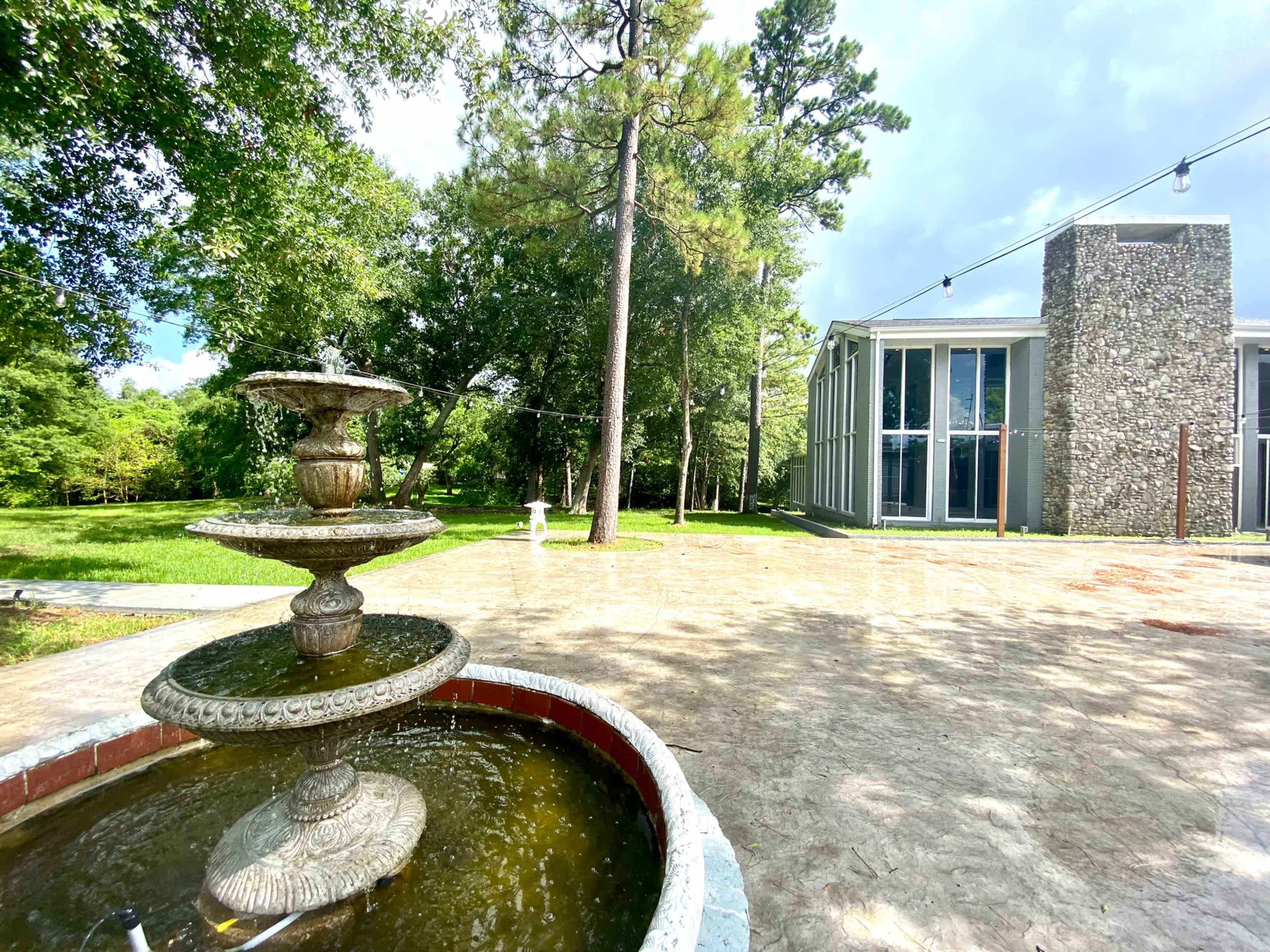 A stone fountain with three tiers sits in the foreground, while a modern glass building and tall trees are visible in the background.