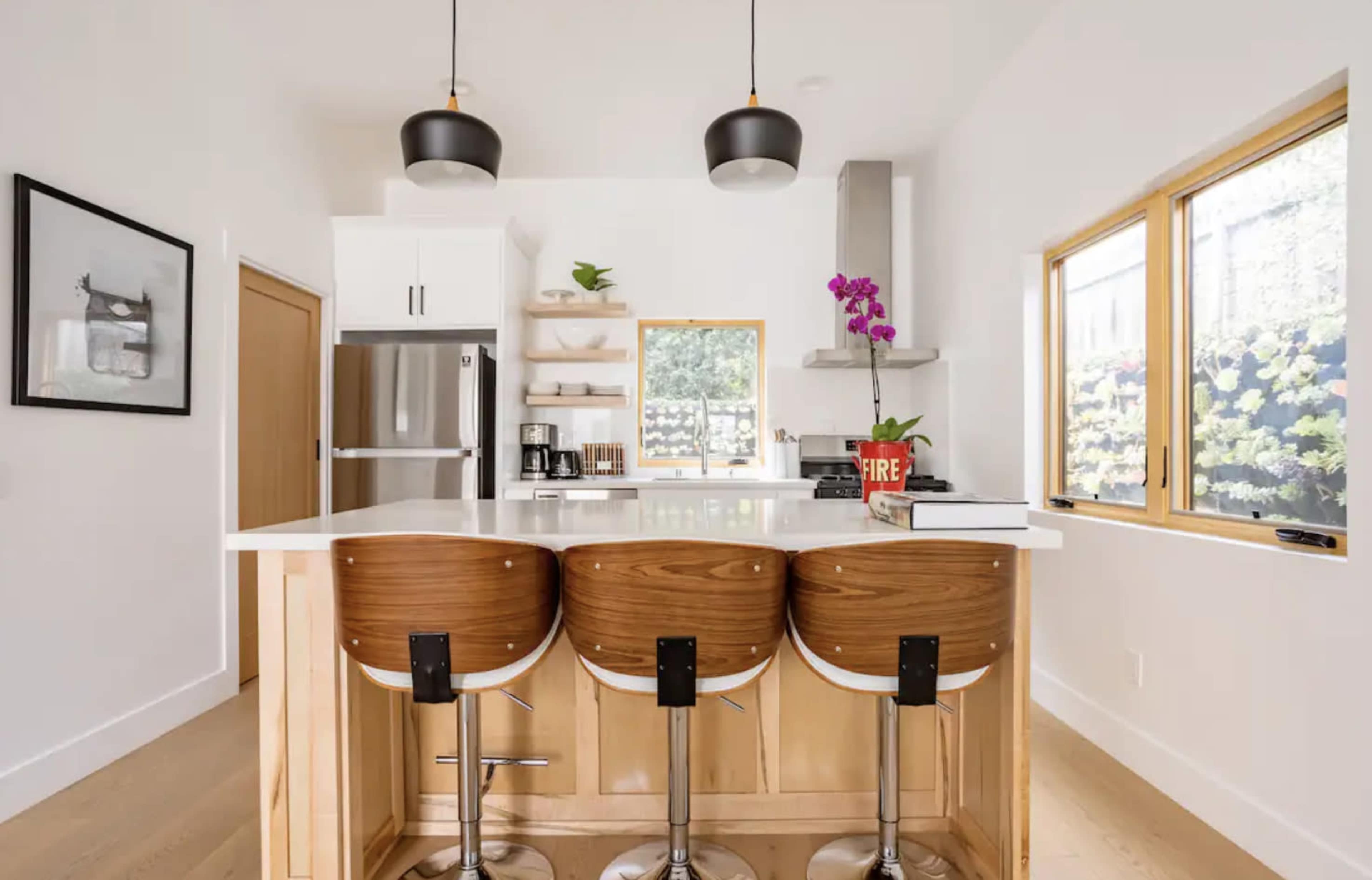 A modern kitchen features a white island with three wooden barstools, stainless steel appliances, and large windows displaying green foliage outside.