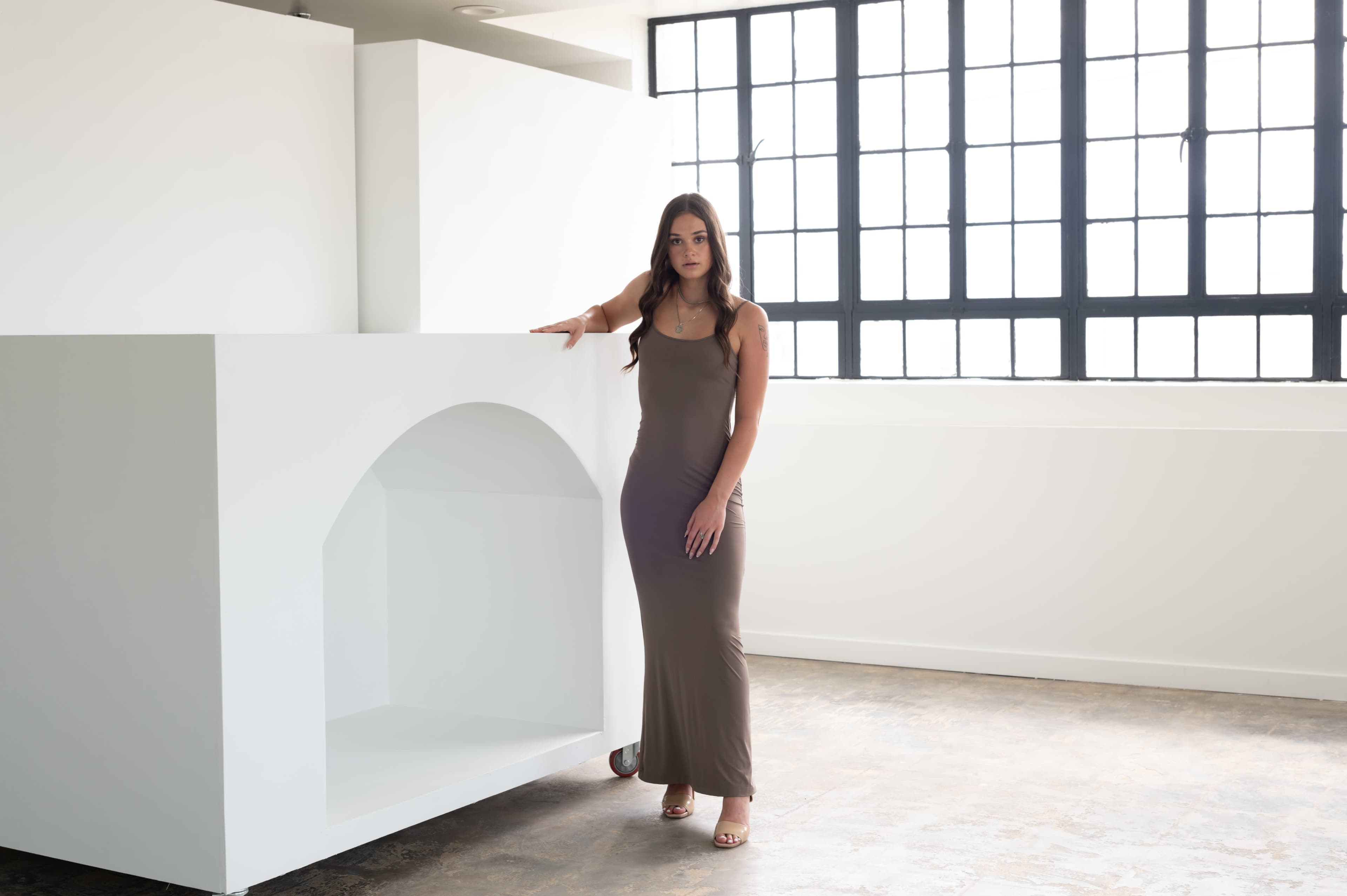 A woman stands next to a large minimalist white structure in a well-lit, modern interior space with large windows.