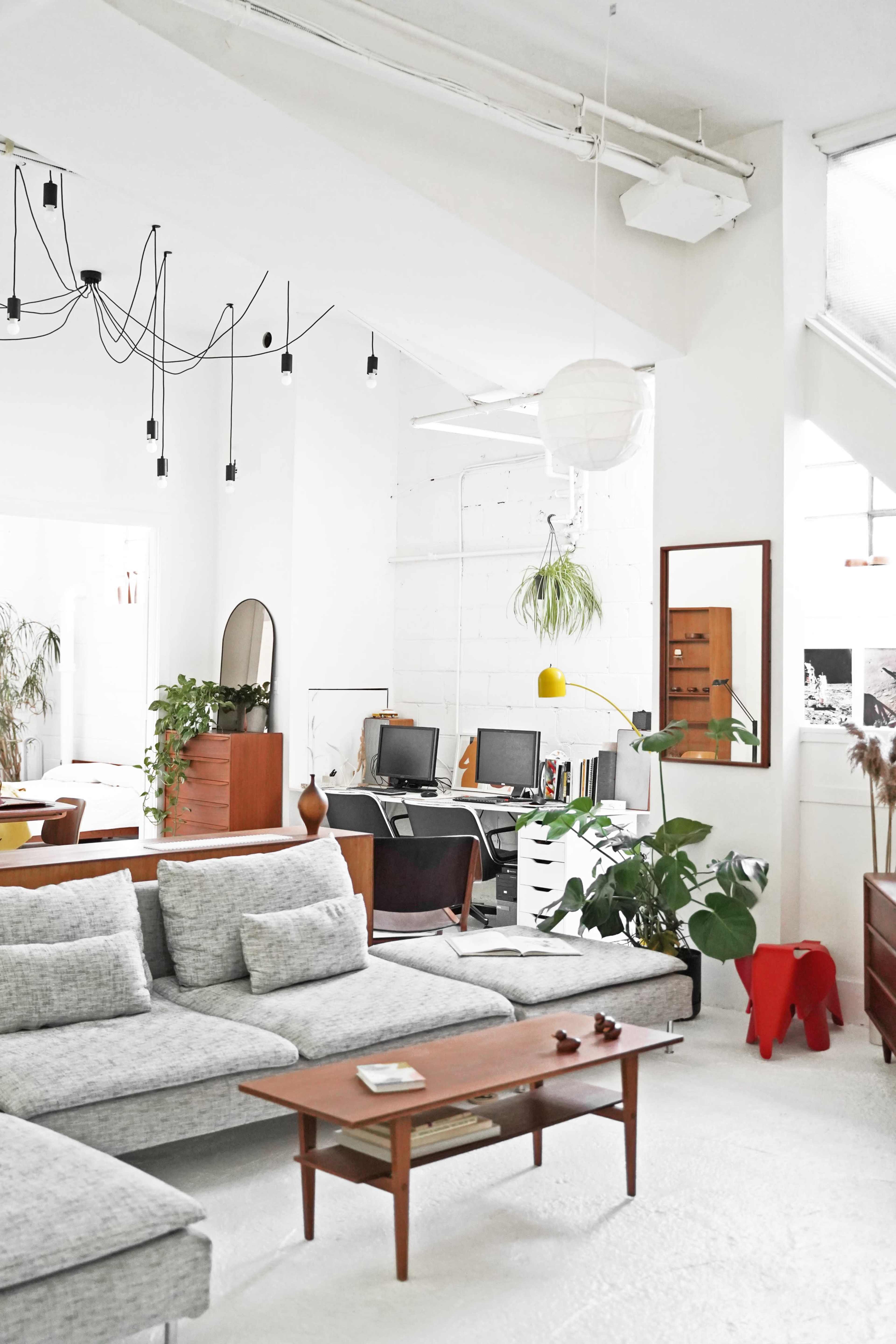 A modern living room features a sectional sofa, a wooden coffee table, plants, and a workspace with two computers in a bright, minimalist space.