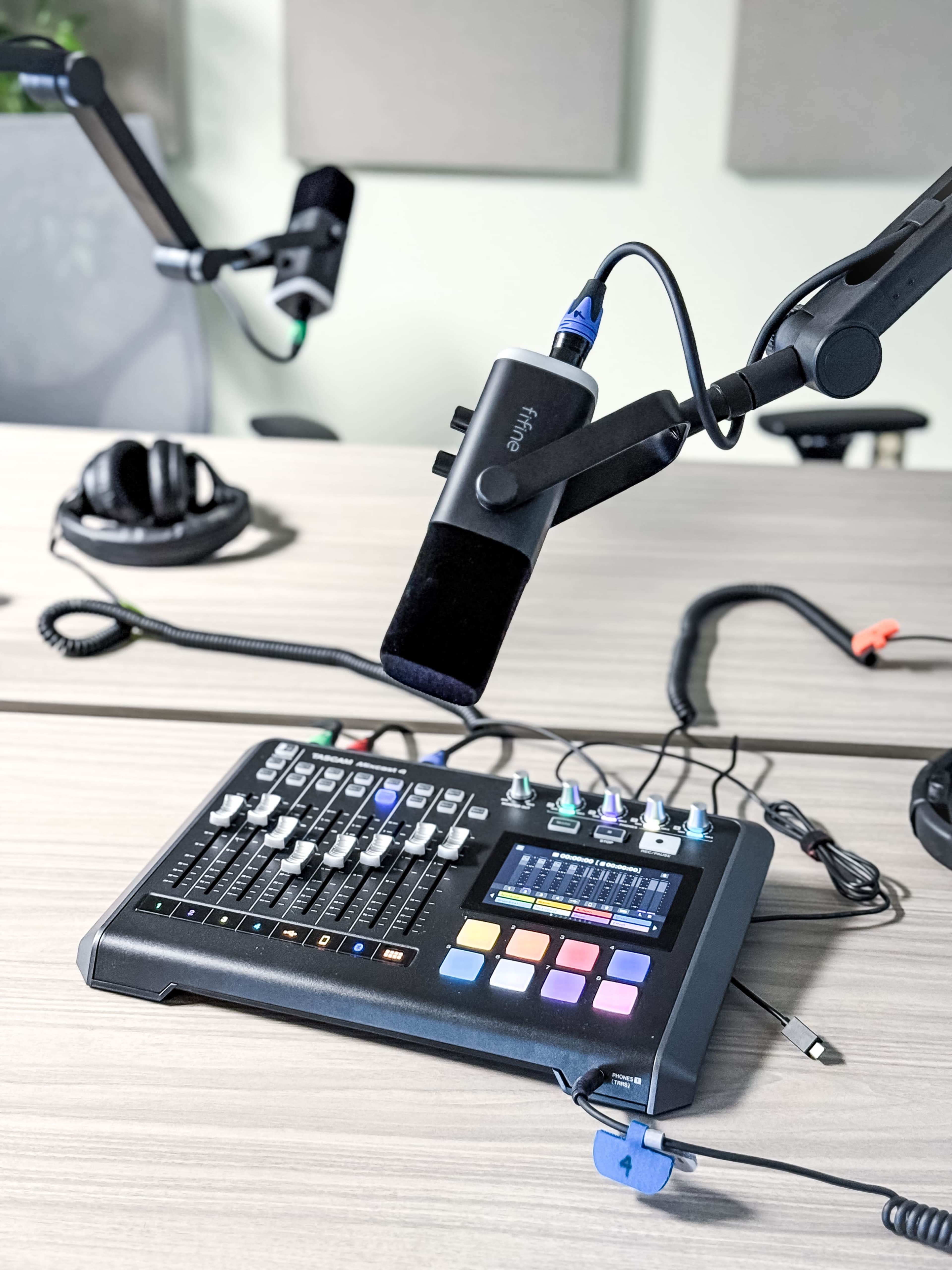 A black audio mixer with colorful buttons and several connected microphones is positioned on a wooden table in a recording studio.