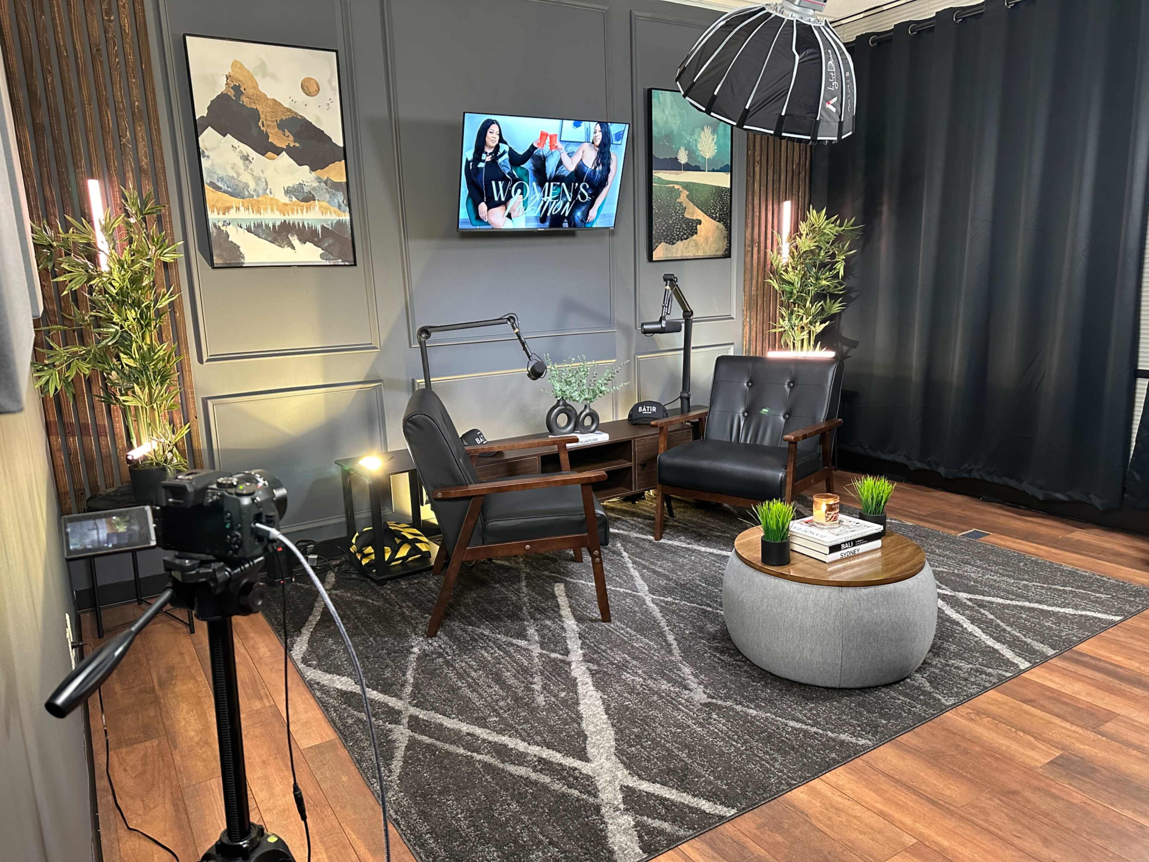 The image shows a modern studio setup with two black chairs, a round table, and a large television screen on the wall, surrounded by decor and lighting equipment.