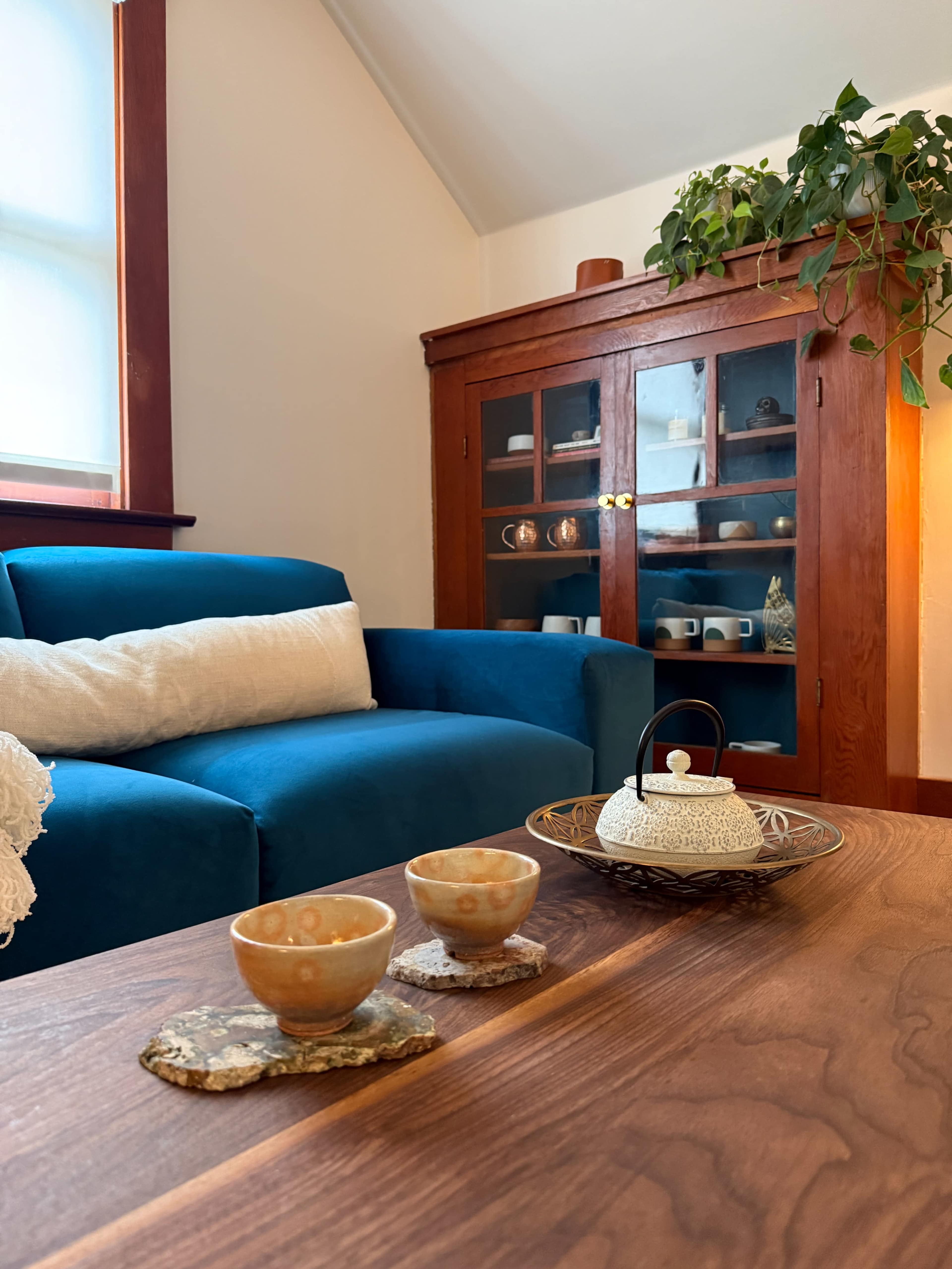 A cozy room features a blue sofa, a wooden table with two tea cups, and a ceramic teapot on a tray, with a glass-front cabinet in the background displaying various cups and decor.