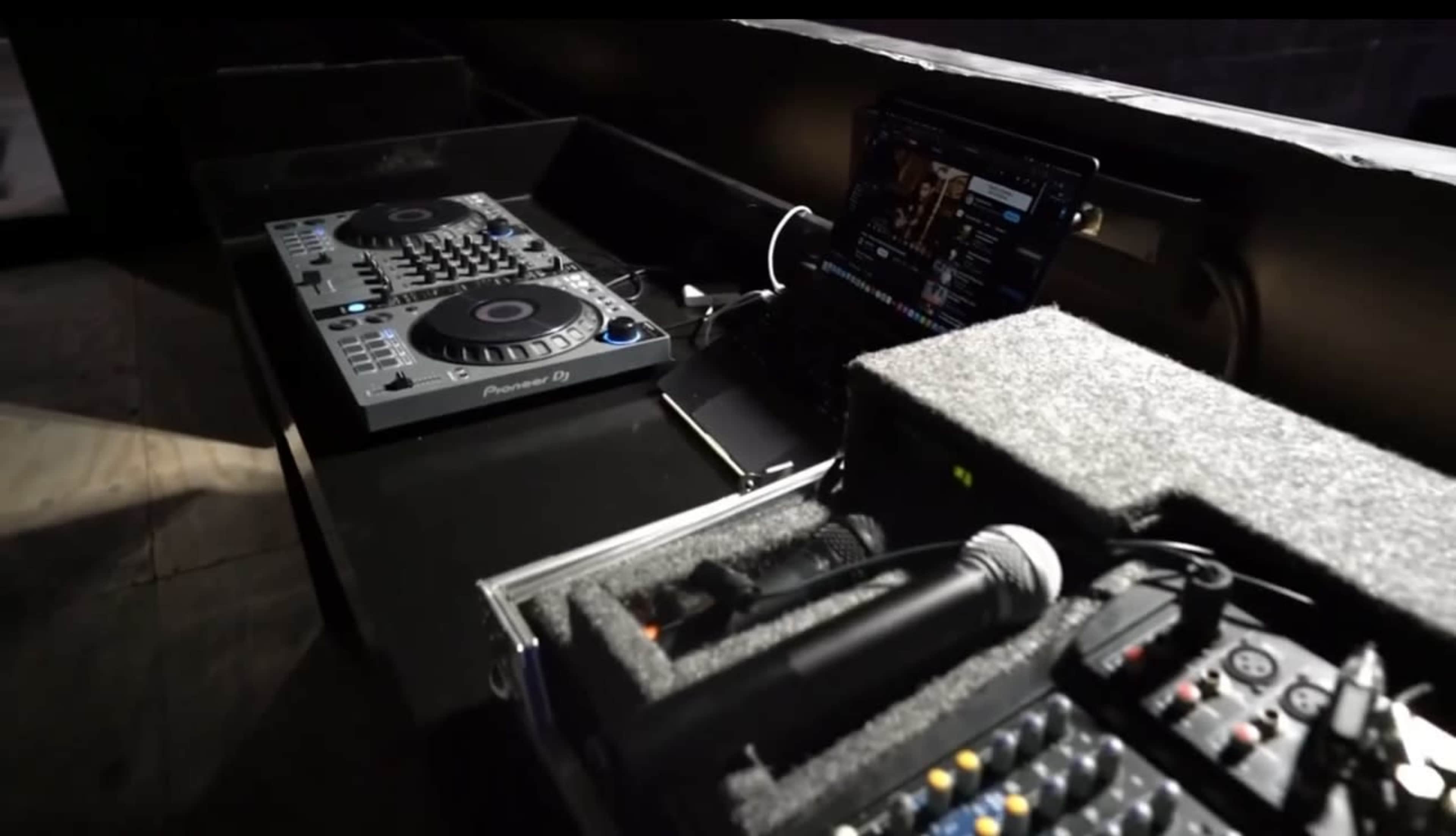 The image shows a DJ setup consisting of two controllers, a laptop on a table, and a microphone within a carrying case.