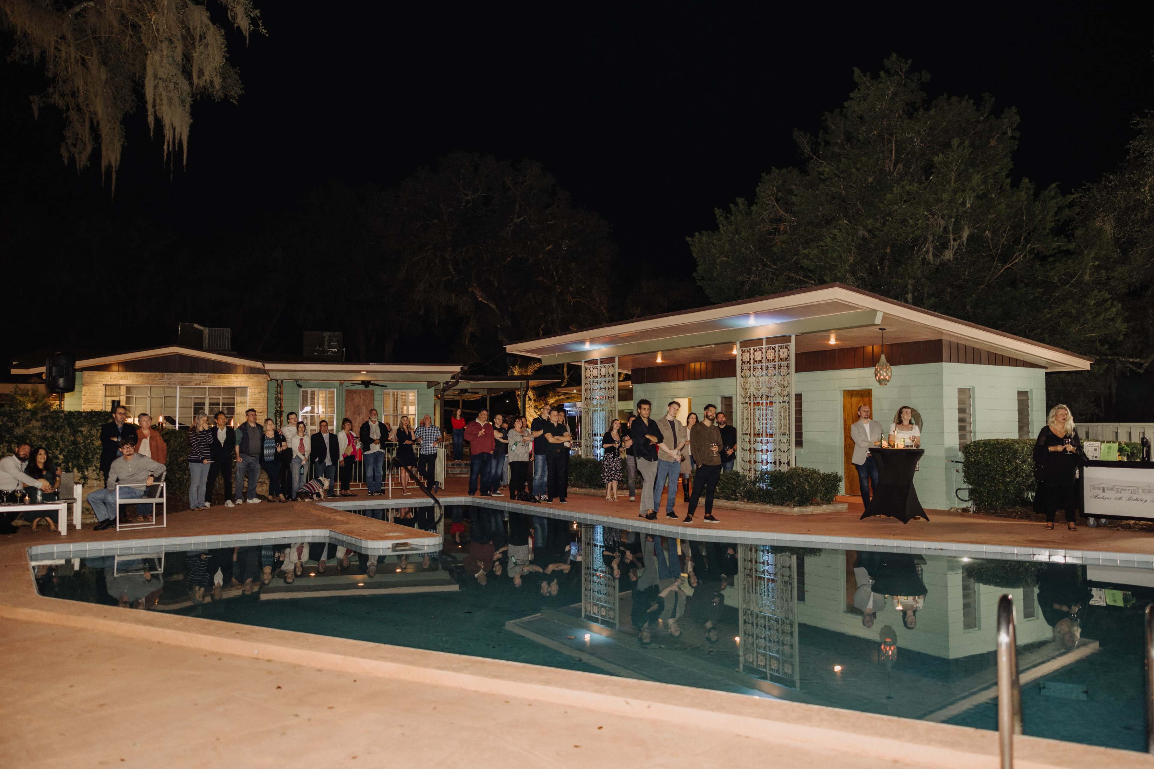 A group of people stands along the edge of a pool at night, with a house and trees visible in the background.