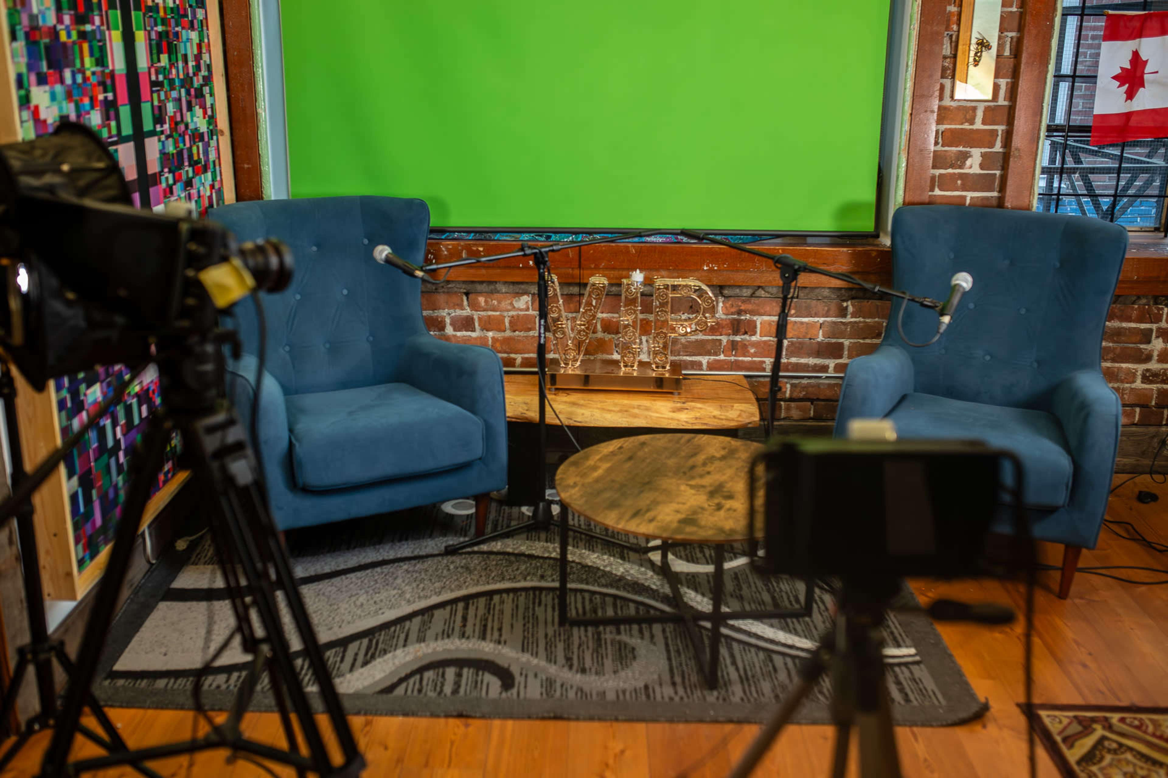 The image shows a small podcasting studio with two blue armchairs, a round wooden table, microphones, and a green screen backdrop.