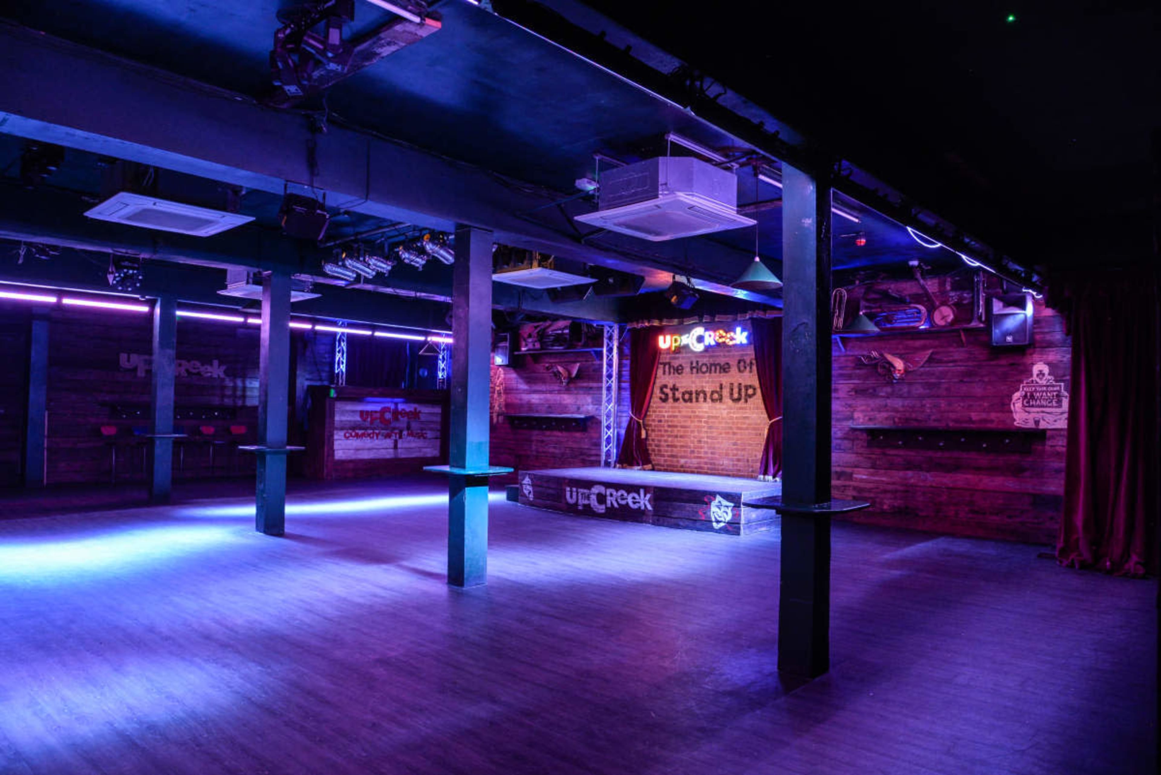 The image shows an empty comedy club interior with wooden walls, stage lighting, and a bar area.