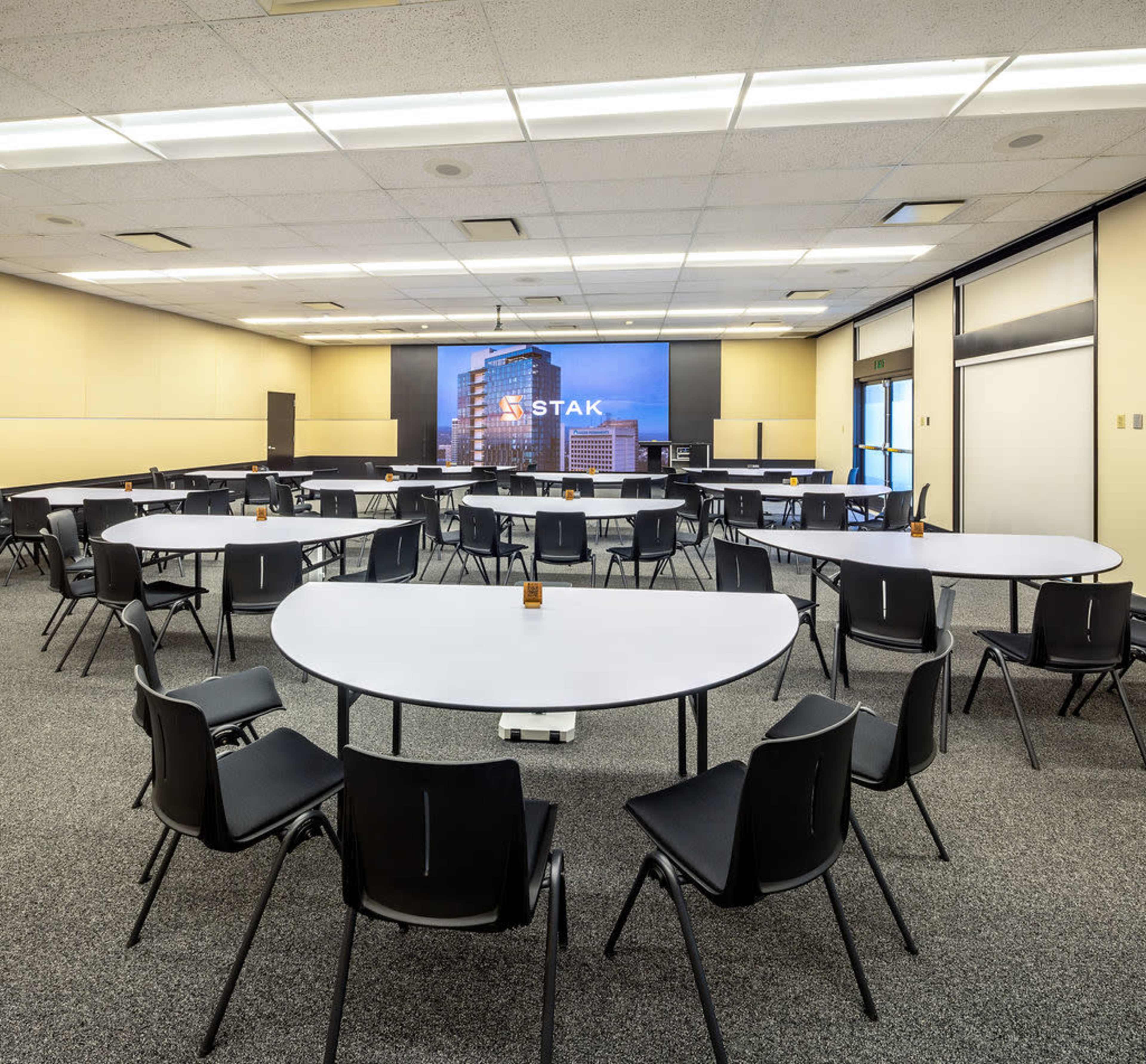 A large meeting room is set up with several round tables and chairs arranged in a semi-circle, featuring a large screen displaying a building image at the front.