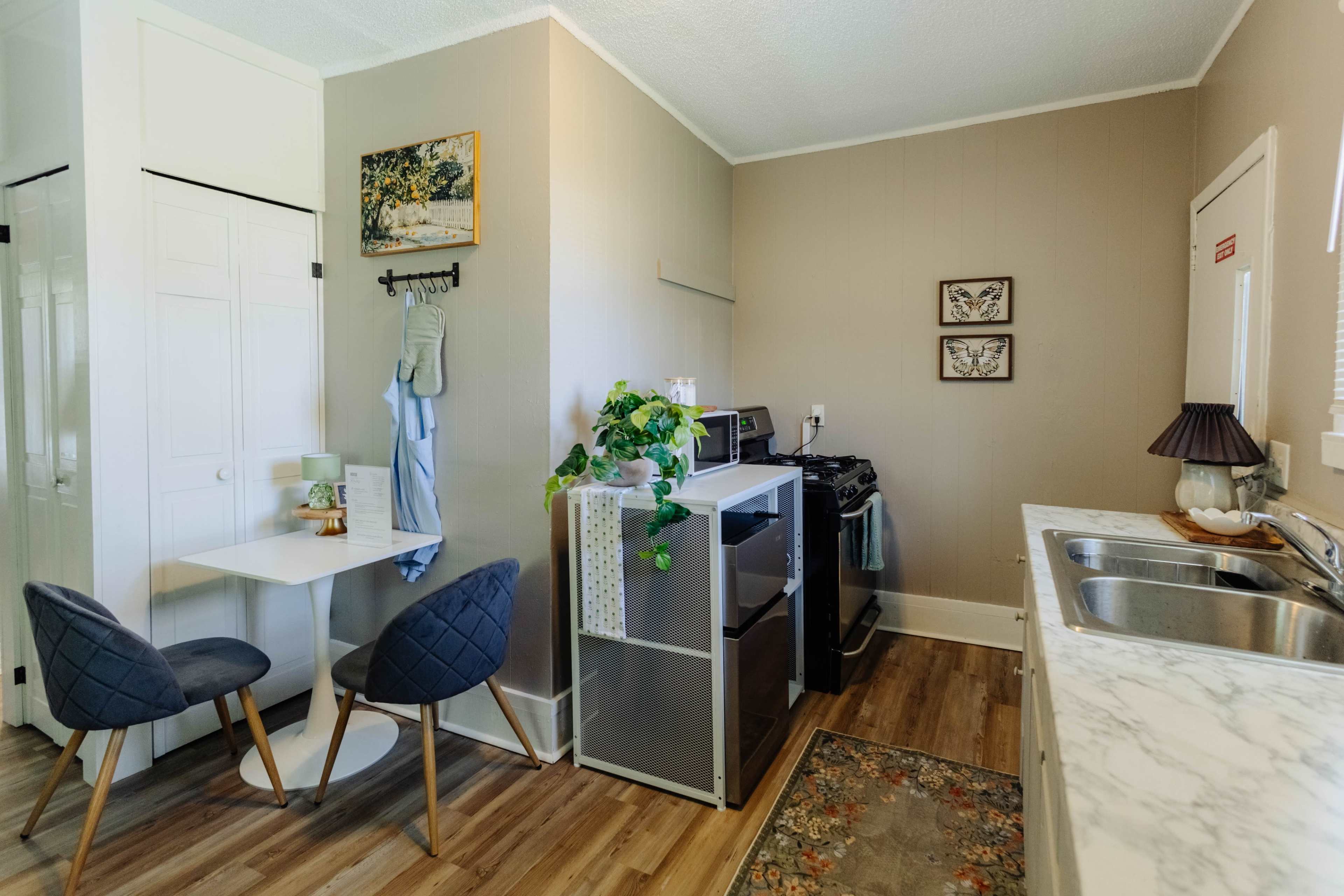 The image shows a compact kitchen area featuring a small dining table with two chairs, a refrigerator, a stove, and a sink with a marble countertop.