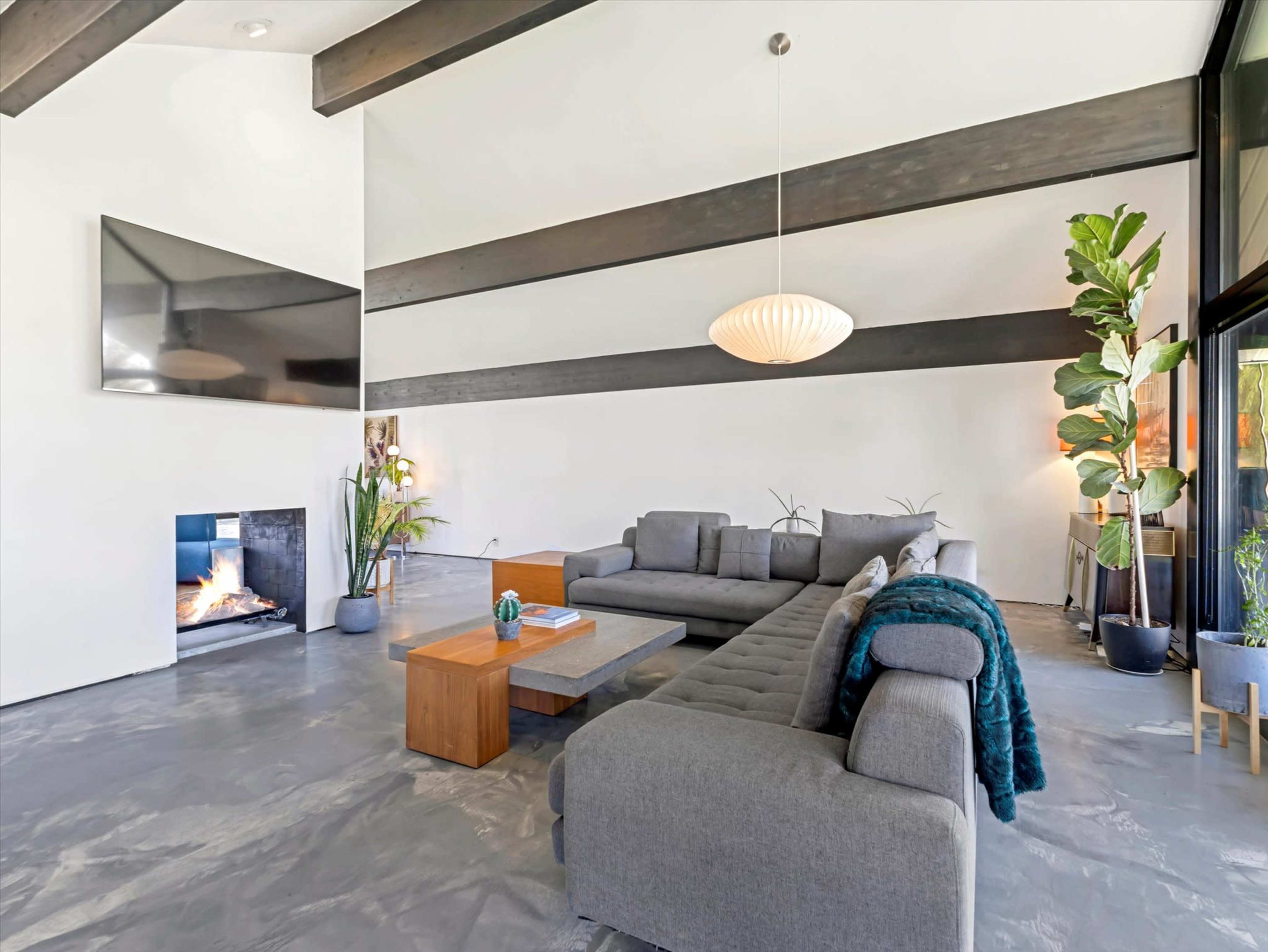 The image shows a modern living room with a large sectional sofa, a coffee table, and a fireplace beneath a flat-screen TV, complemented by indoor plants and a high ceiling with exposed beams.