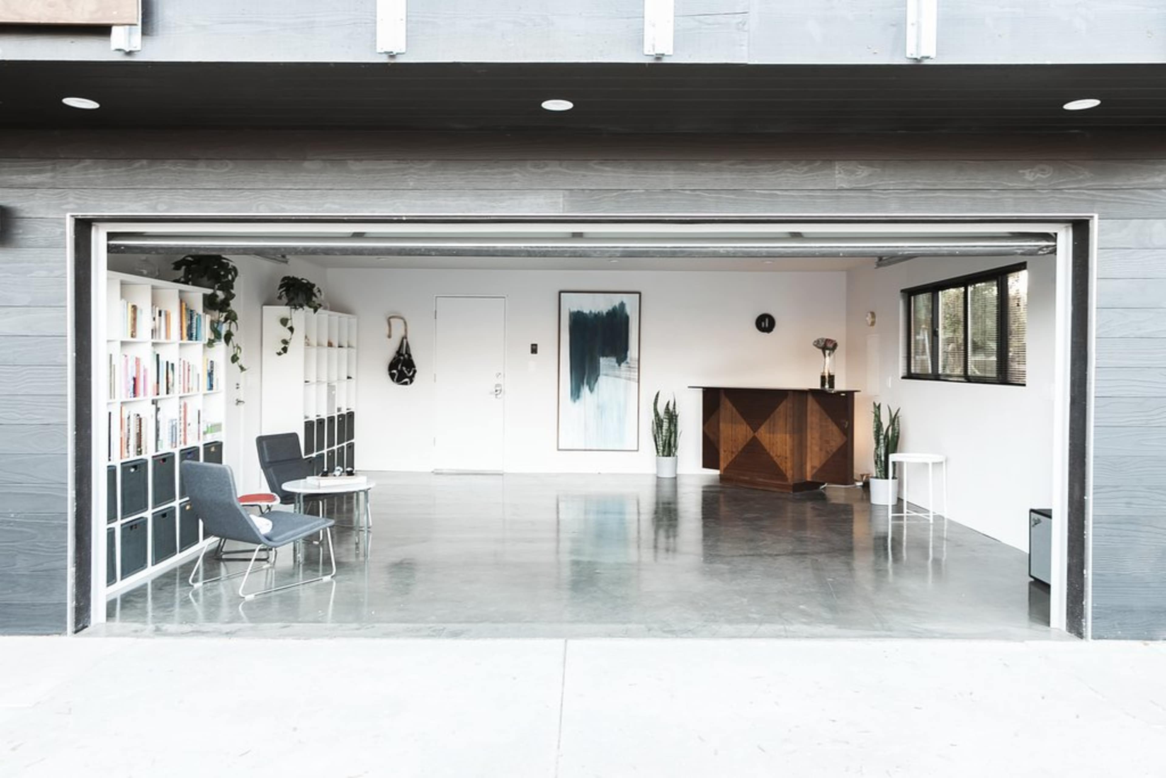 A spacious interior features a garage door open to a minimalist living area with bookshelves, a bar cabinet, and potted plants.