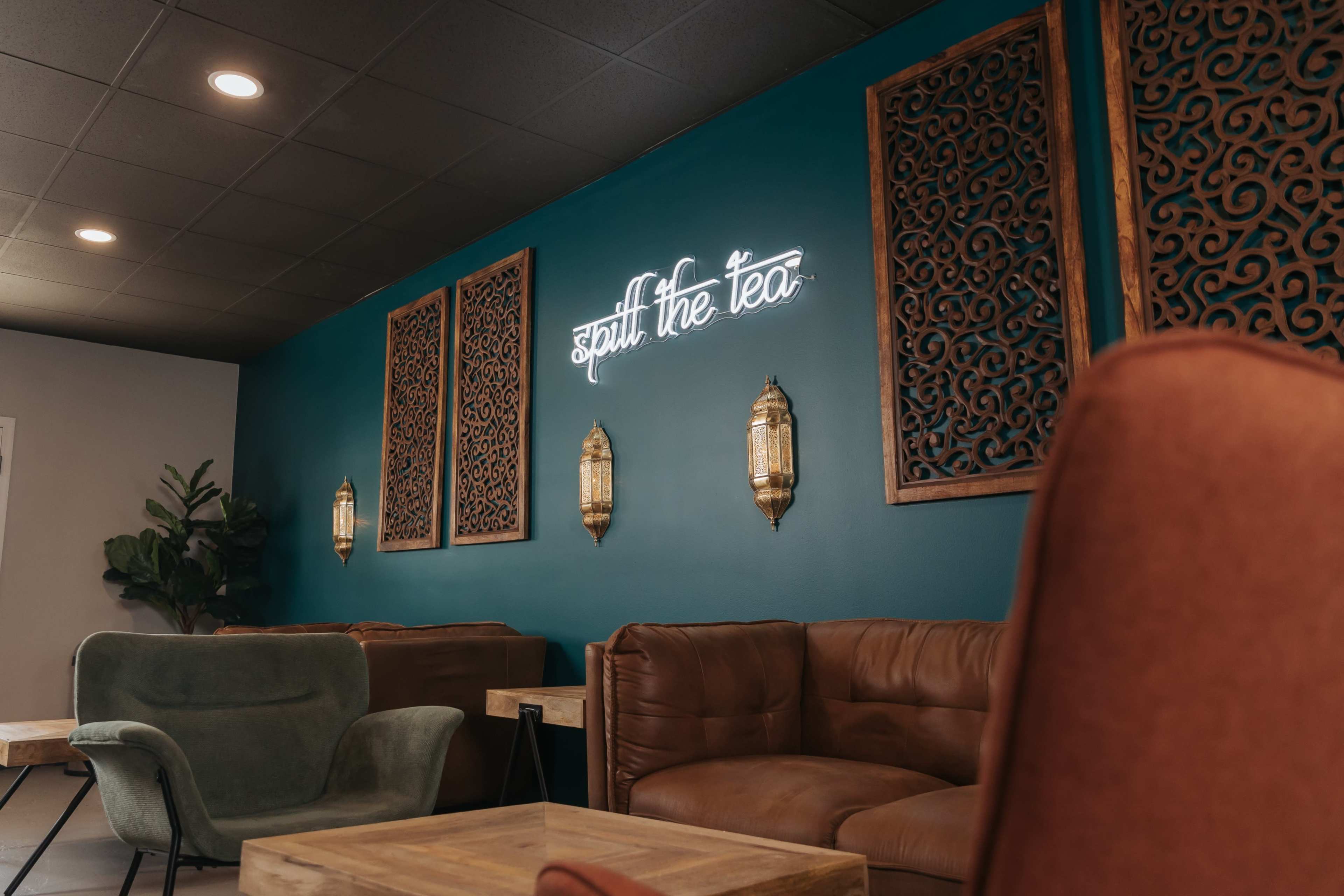 The interior of a cozy lounge features a teal wall with decorative panels and a neon sign that reads "Spill the Tea."