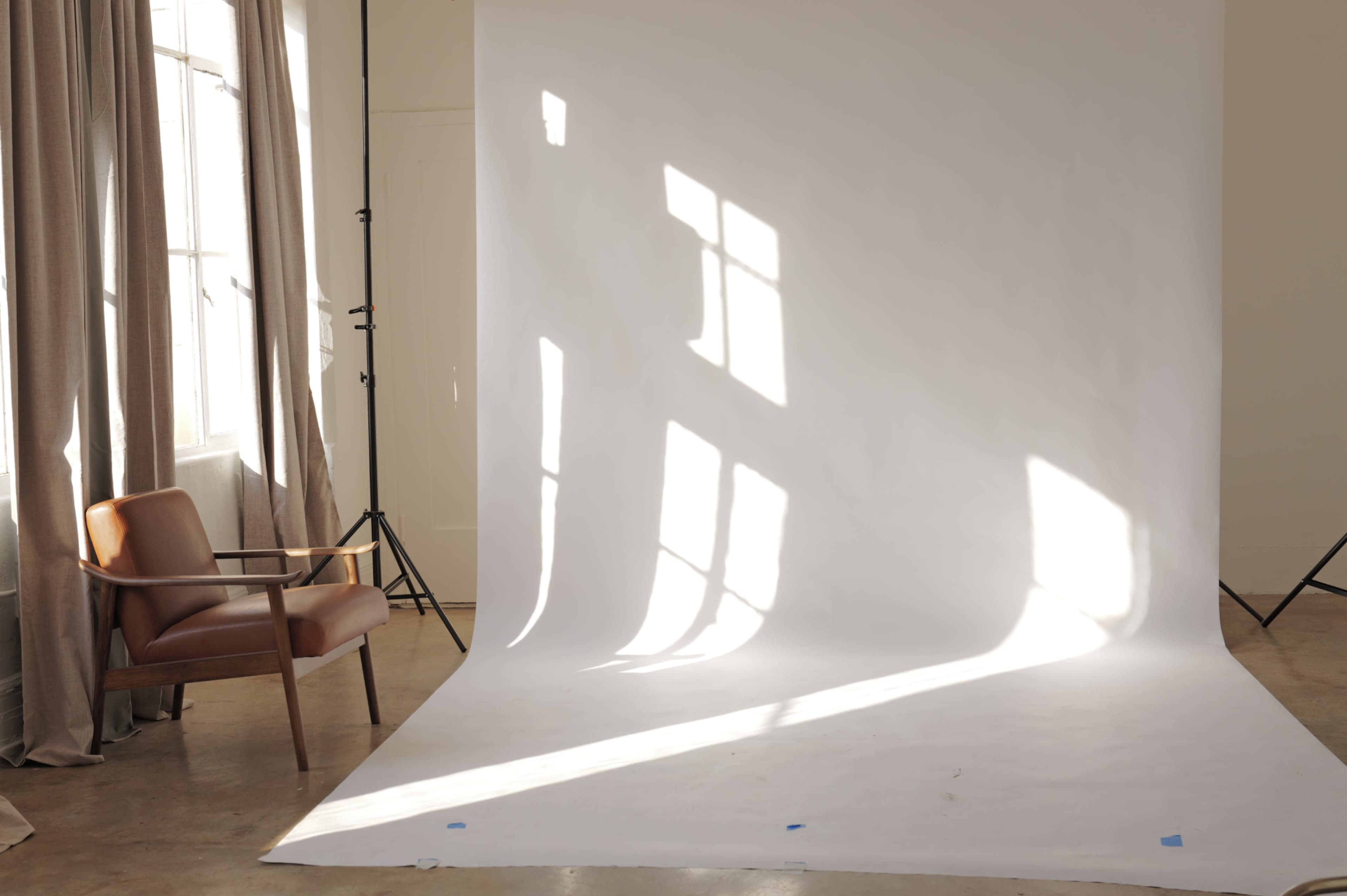 The image shows a spacious photography studio with a white backdrop, a wooden chair, and sunlight casting shadows from windows onto the floor.