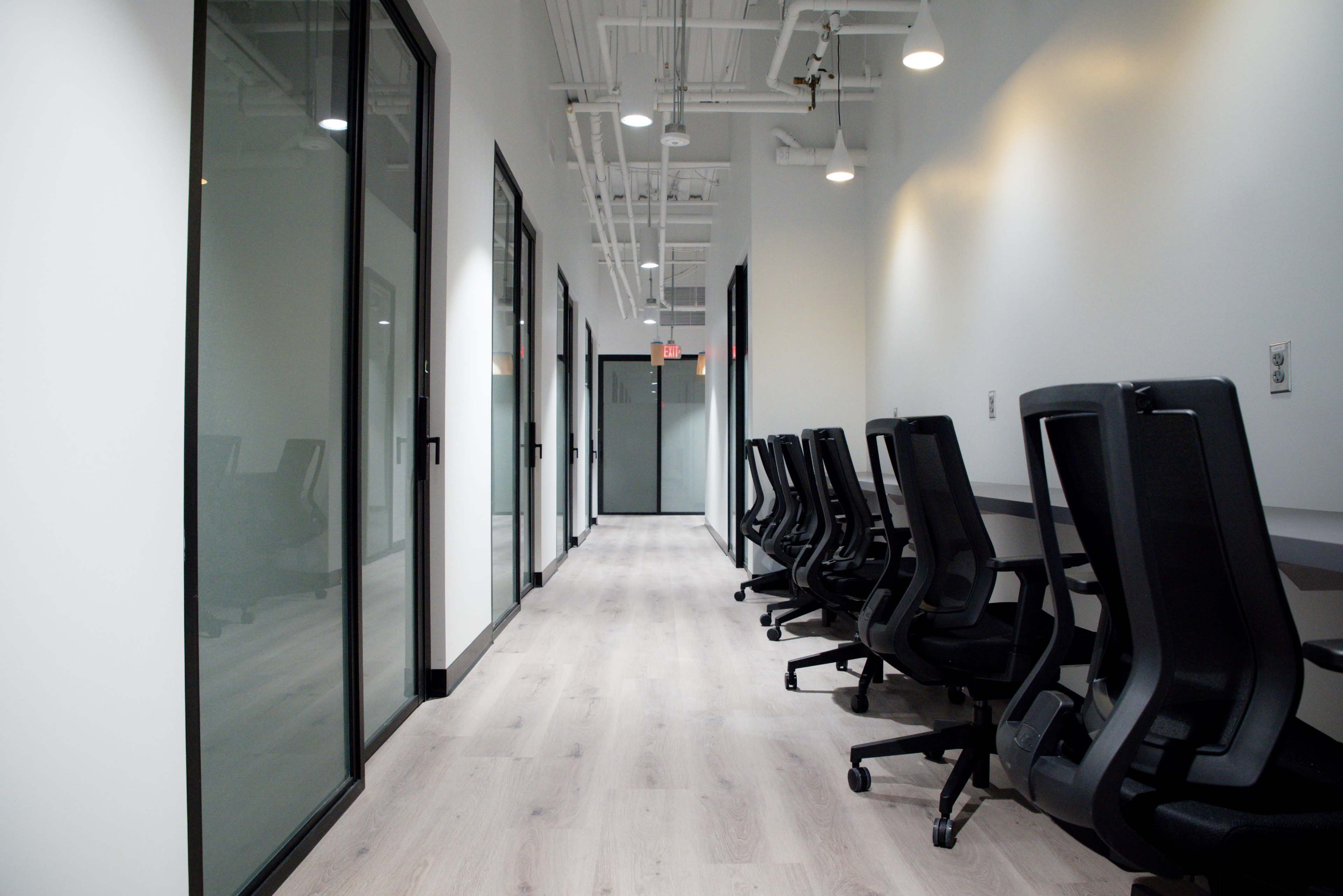 The image shows a modern office hallway with glass-walled meeting rooms and a row of black office chairs along one side.