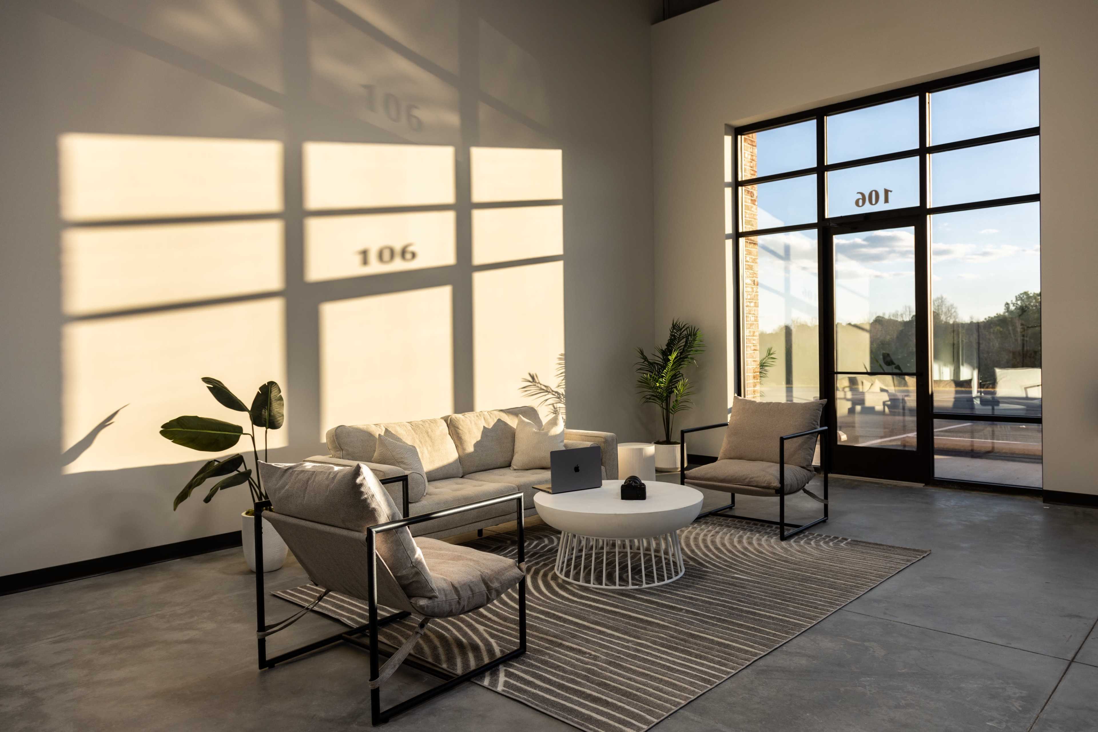 A modern living space features a beige sofa, two chairs, and a round coffee table set on a striped rug, with sunlight casting geometric shadows on the wall.