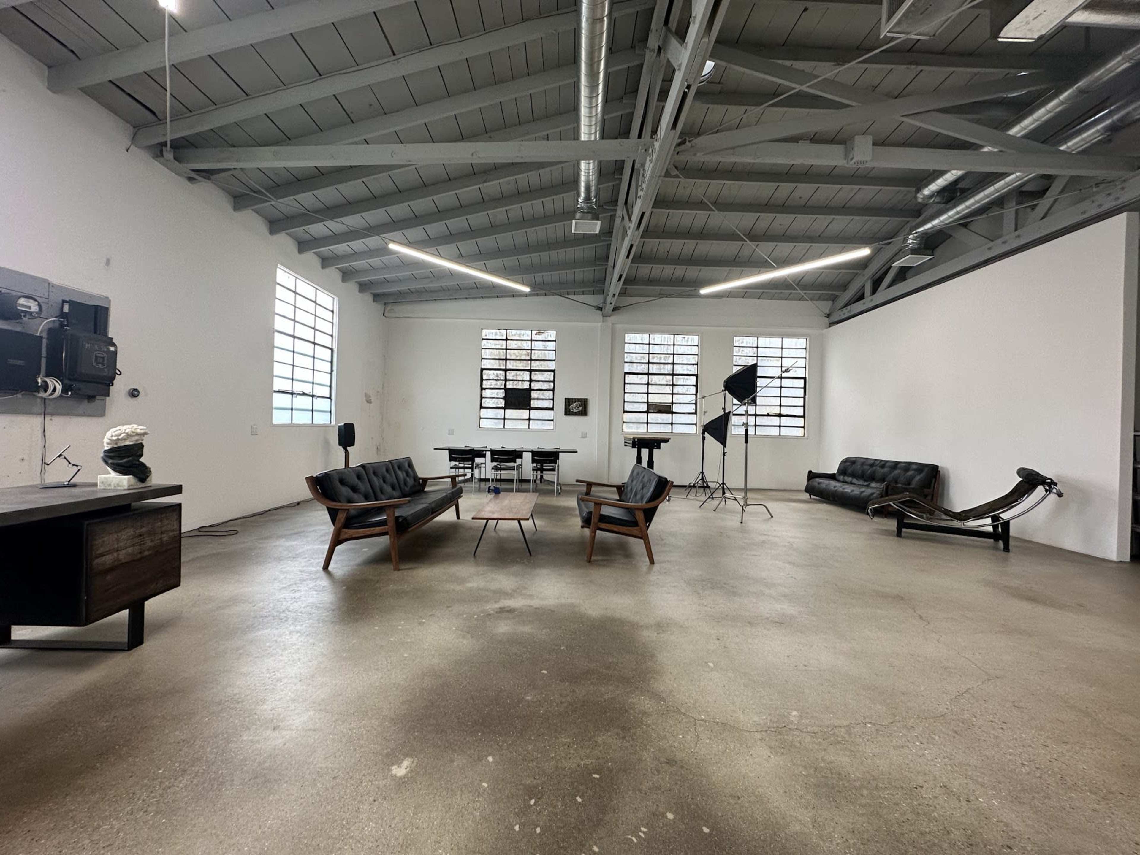 A spacious, industrial-style room with high ceilings, featuring modern furniture including a seating area, dining table, and several large windows.