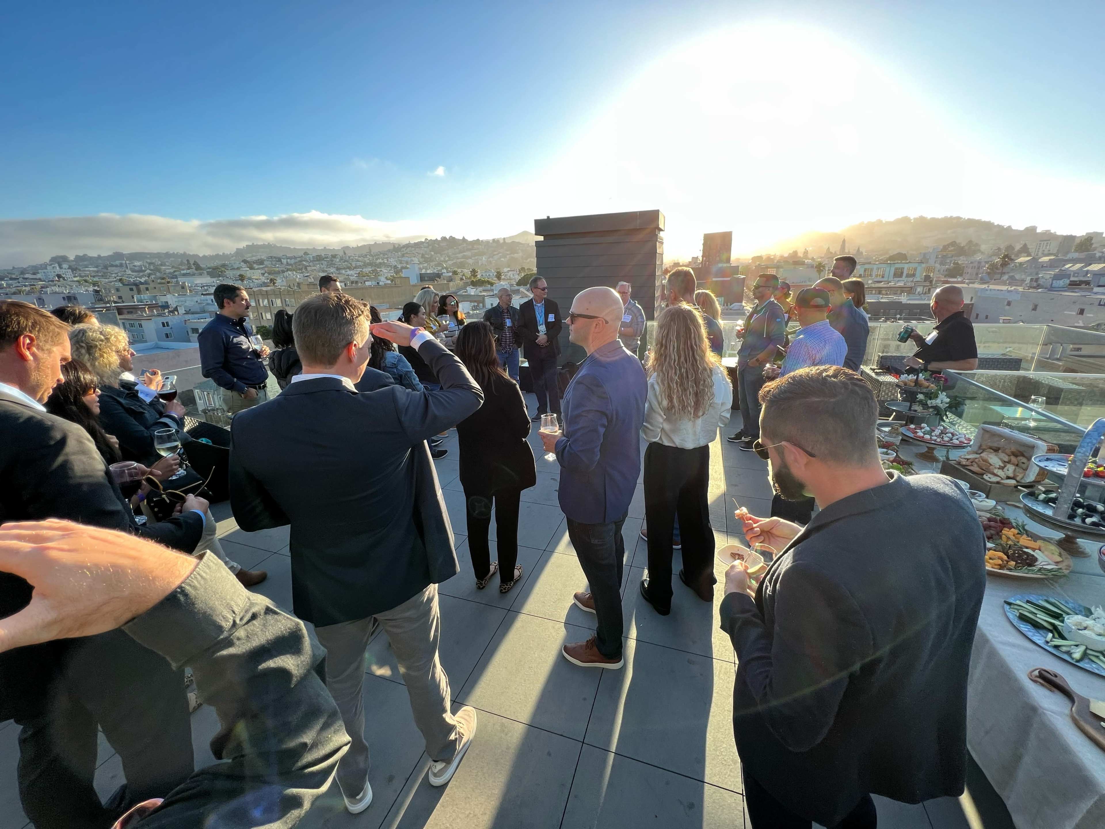 A group of people gathers for a networking event on a rooftop with a city skyline and sunset in the background.