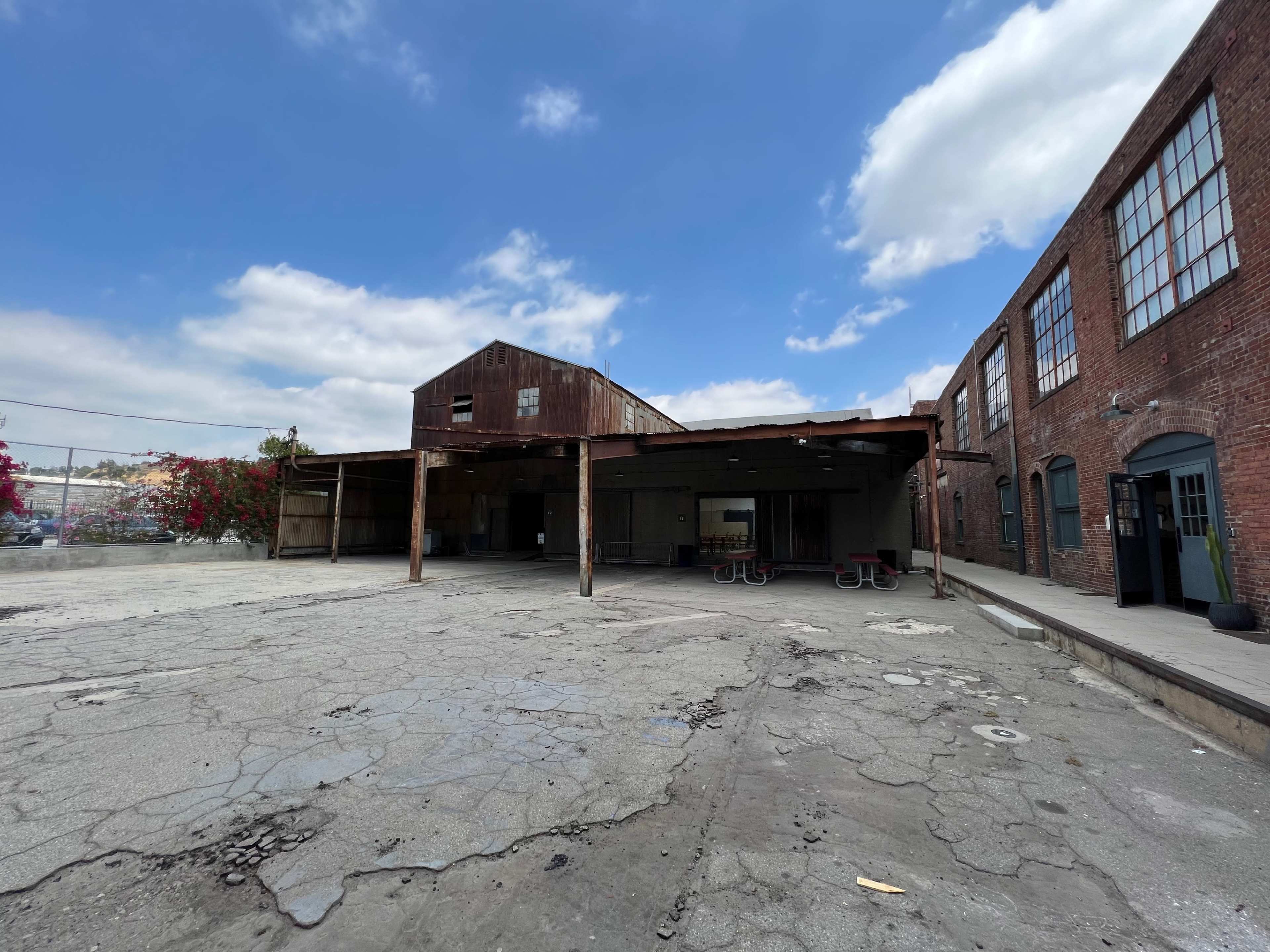 An industrial building with a rusty exterior and a covered patio area sits in an open, cracked concrete space under a partly cloudy sky.