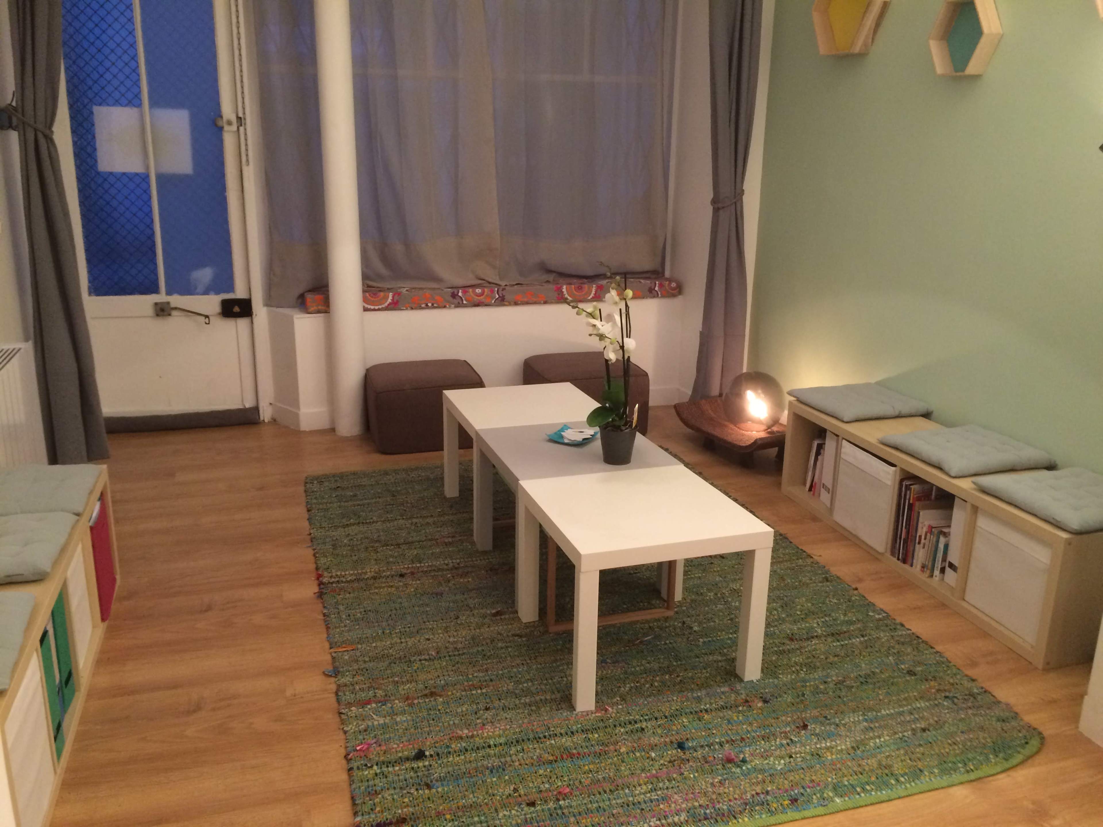 The image shows a cozy room with a wooden floor, featuring a white table at the center, two benches with cushions on either side, and a green accent wall with a decorative plant on the table.
