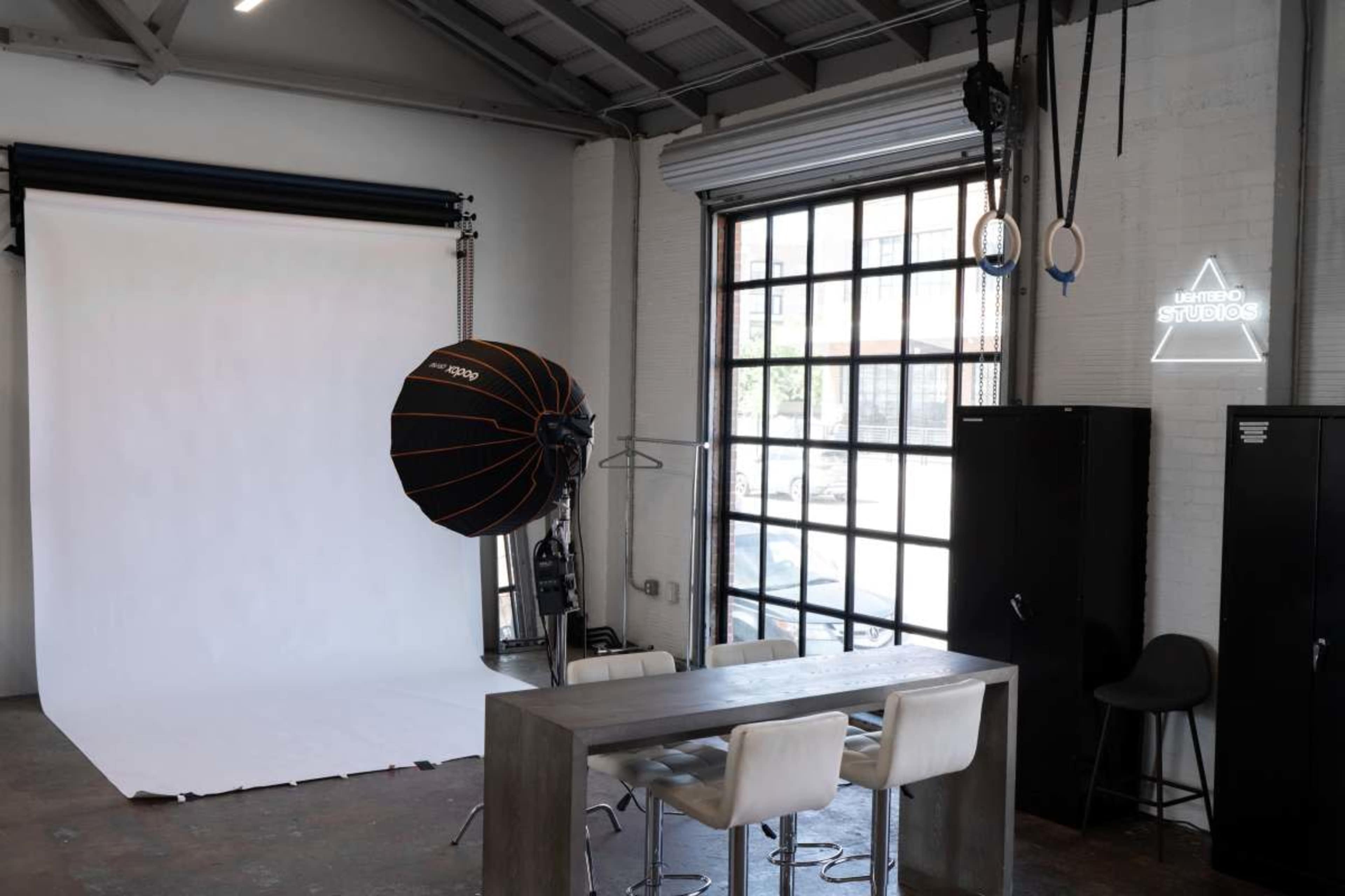 The image shows a photography studio with a white backdrop, a large light source, a table with chairs, and shelving units against a window.