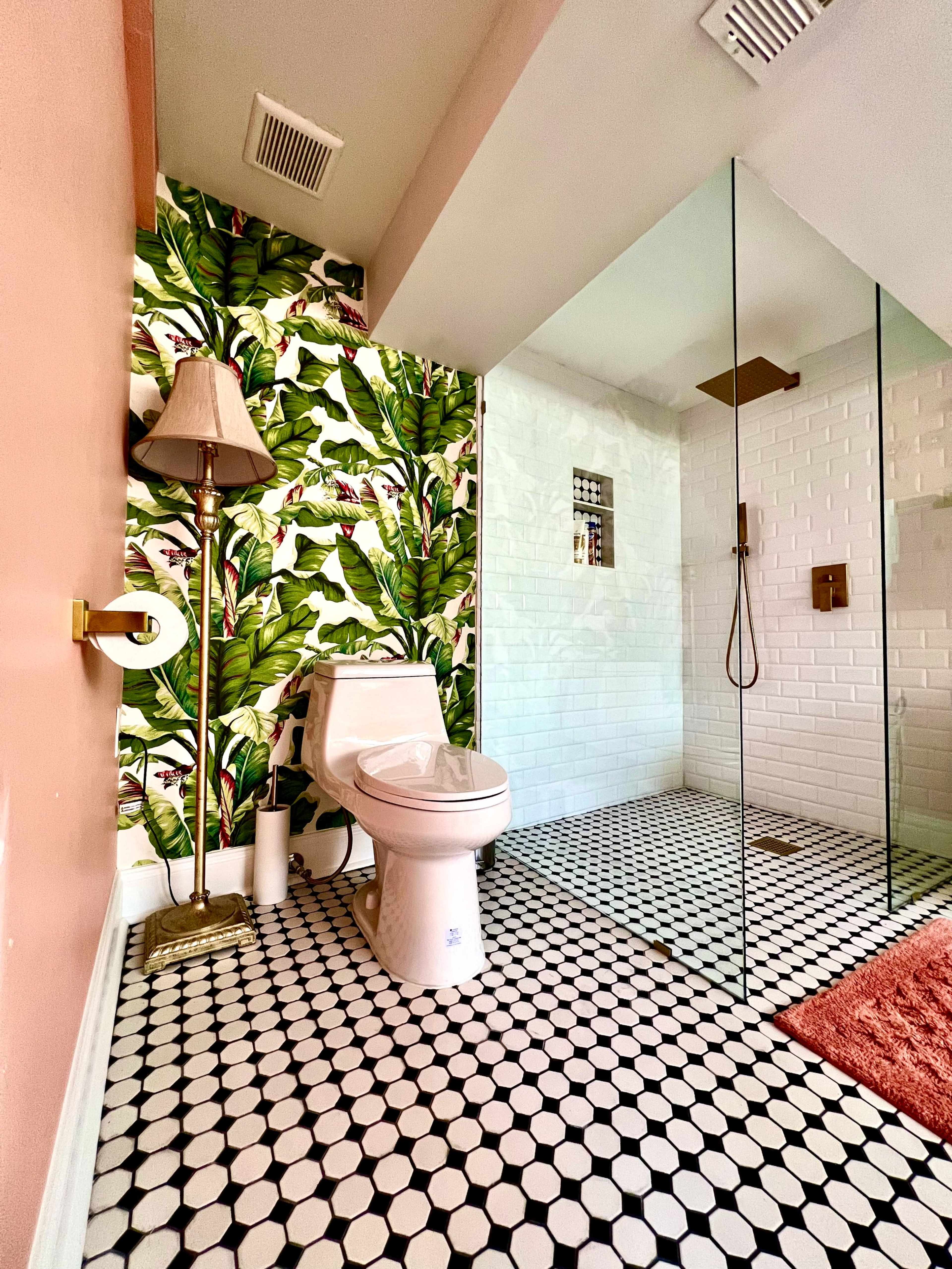 The image features a bathroom with a modern design, showcasing a toilet, a walk-in shower with glass walls, and a vibrant tropical leaf-patterned wallpaper on one wall.