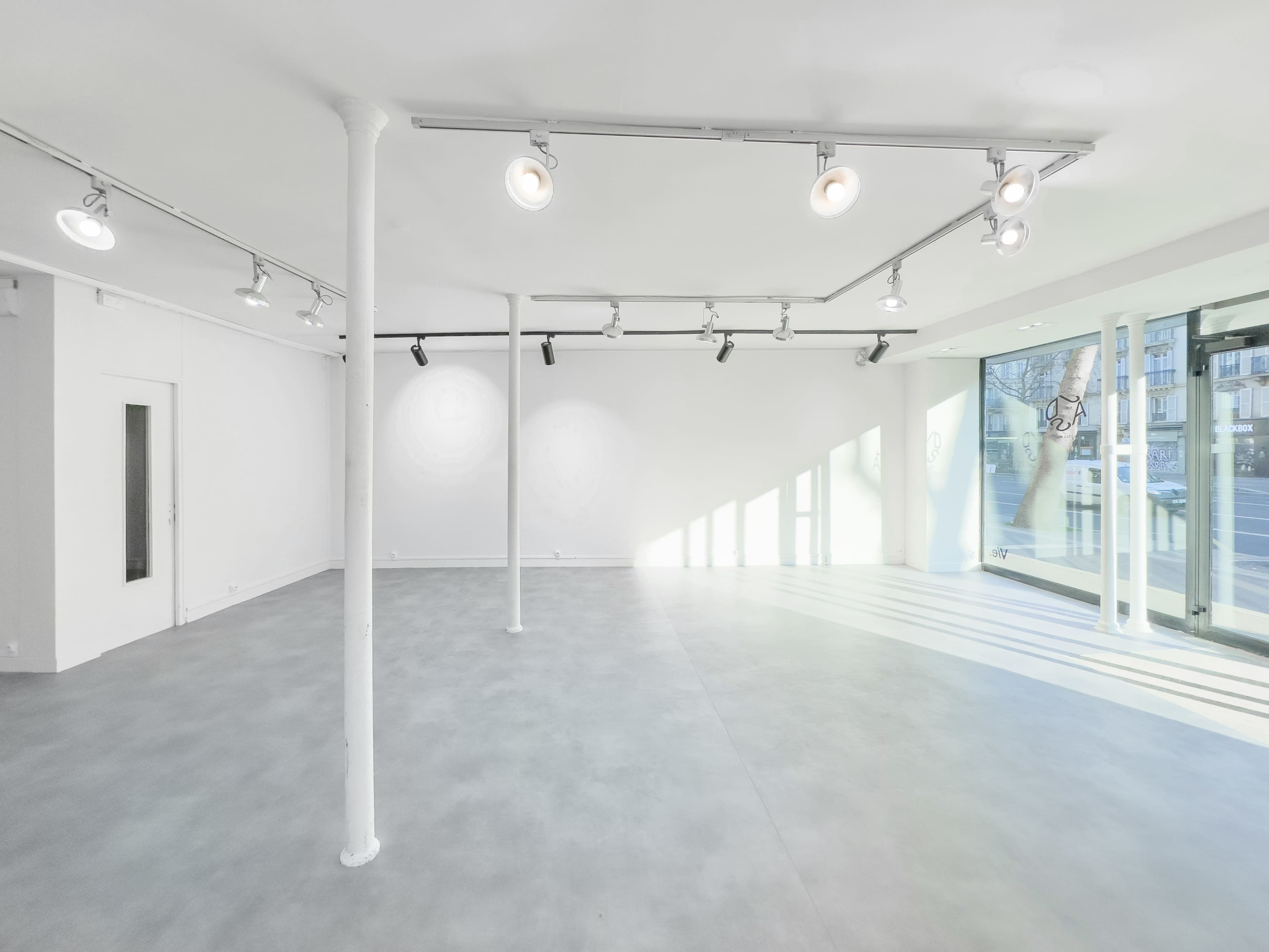 The image shows a spacious, empty room with white walls, a gray floor, and track lighting, featuring large windows that let in natural light.