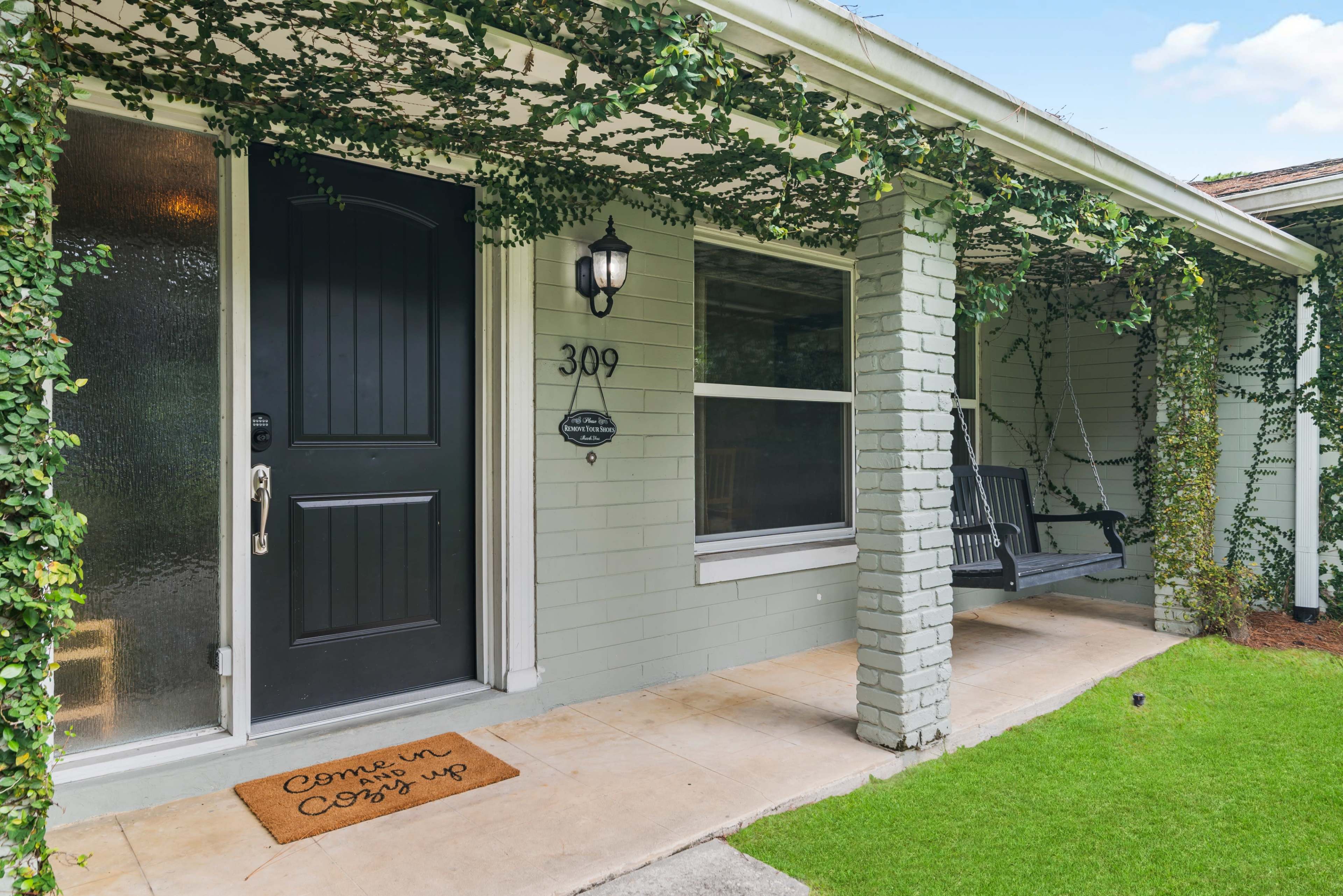 A house entrance with a black front door, a welcome mat, and a porch swing, surrounded by green vines.