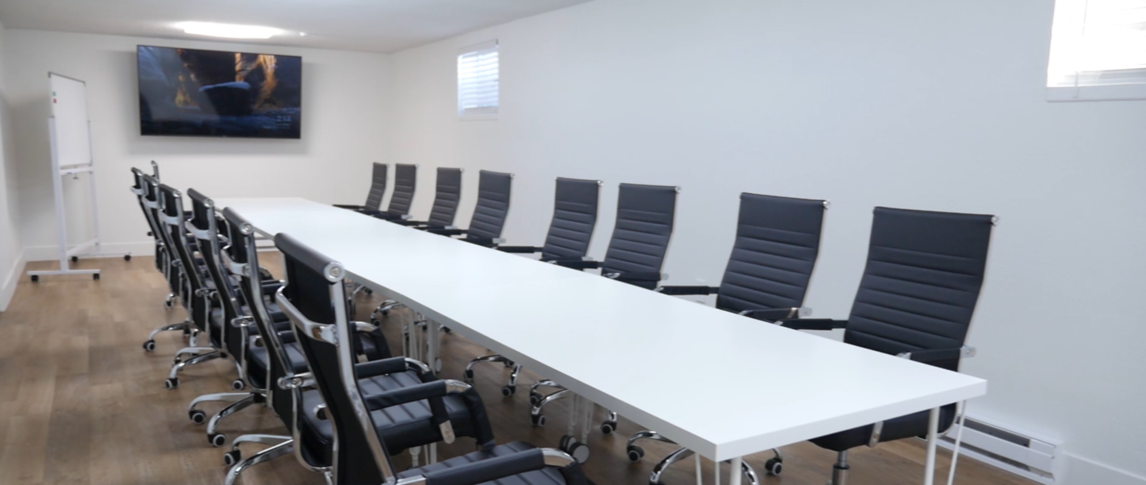 A long, white conference table is flanked by several black office chairs in a bright, minimalist meeting room.