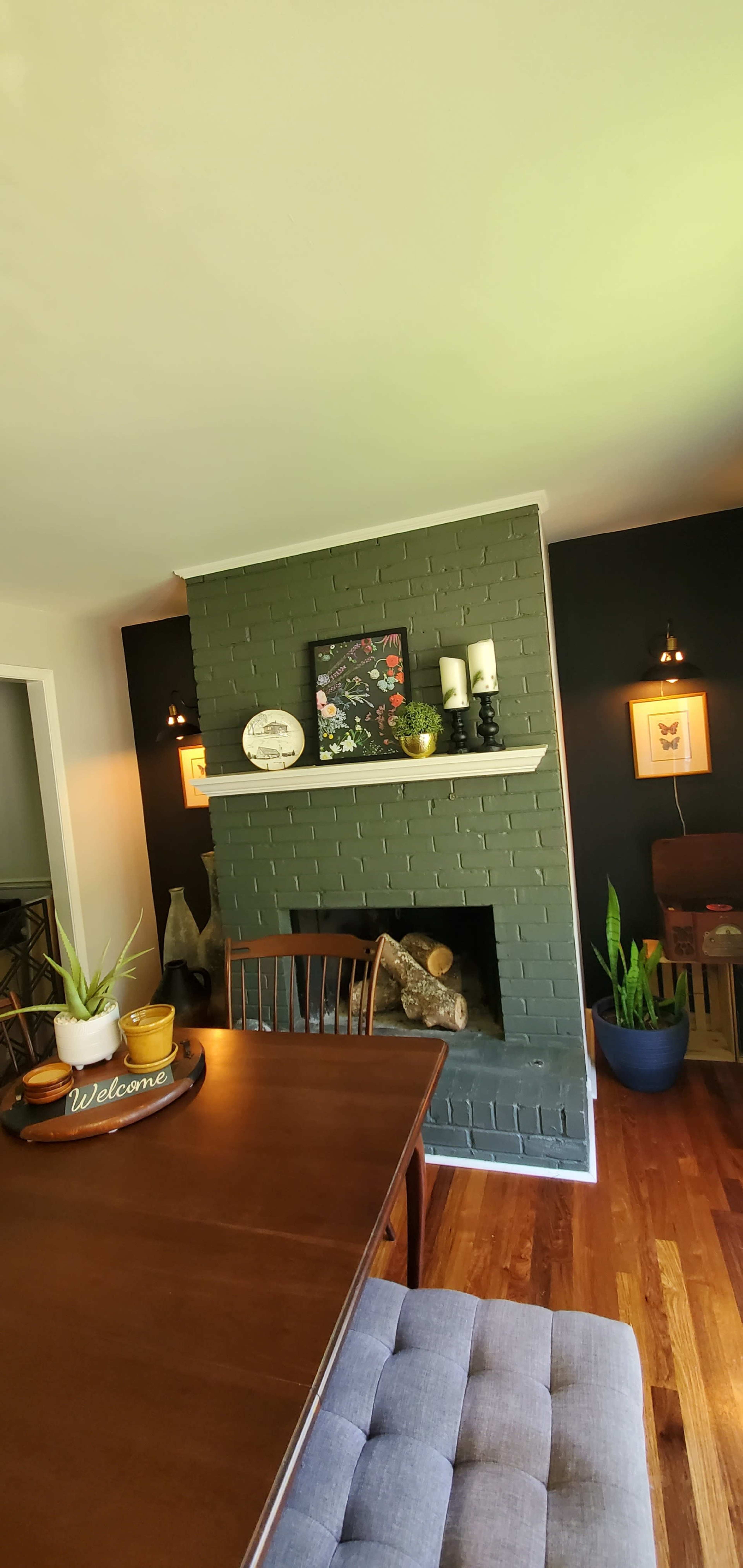 A dining area with a wooden table, a brick fireplace with candles and decorative items, and potted plants in the corners against dark-colored walls.