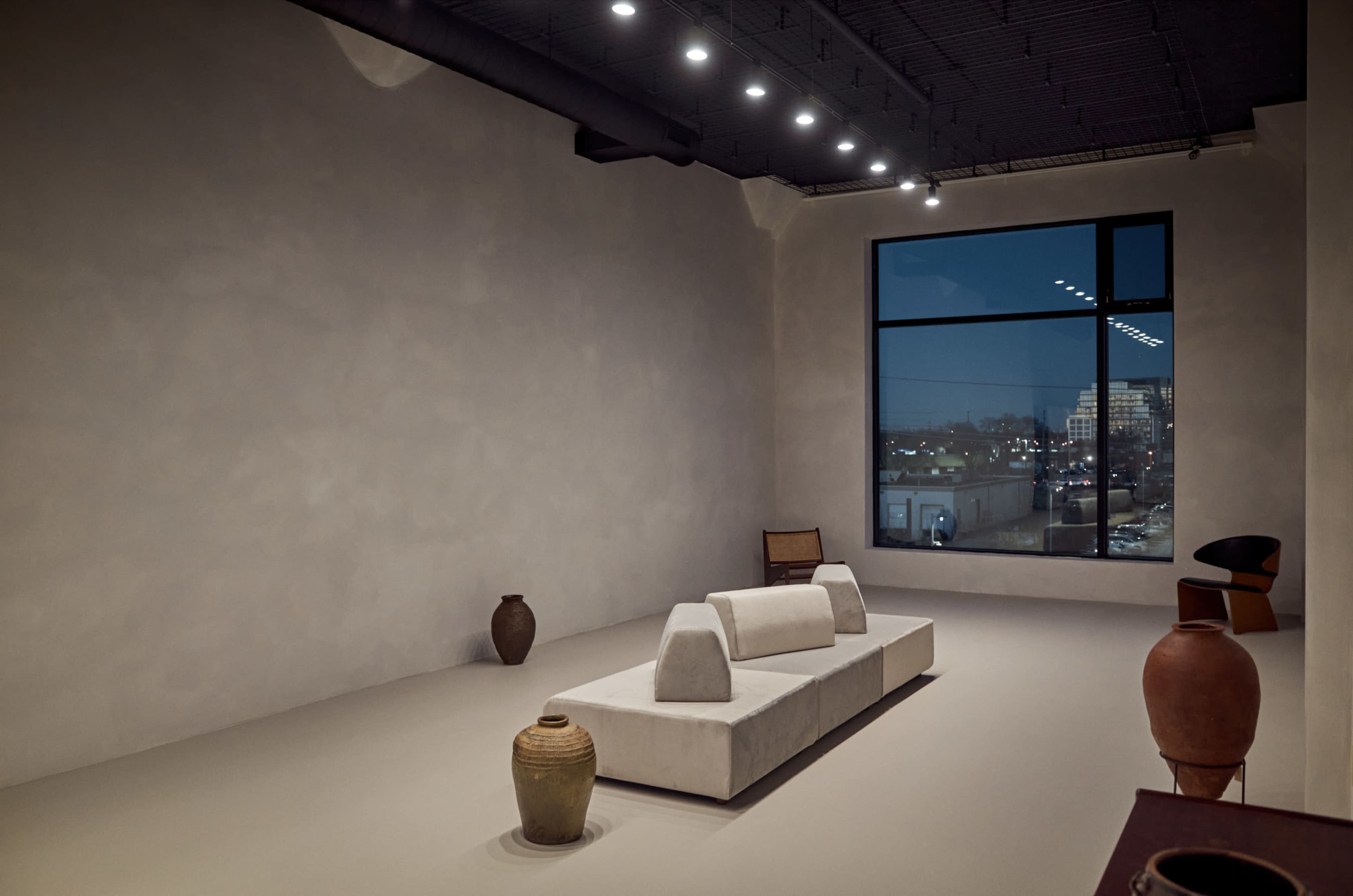 A minimalist room features a central sofa, pottery, and large windows showing a city skyline at dusk.