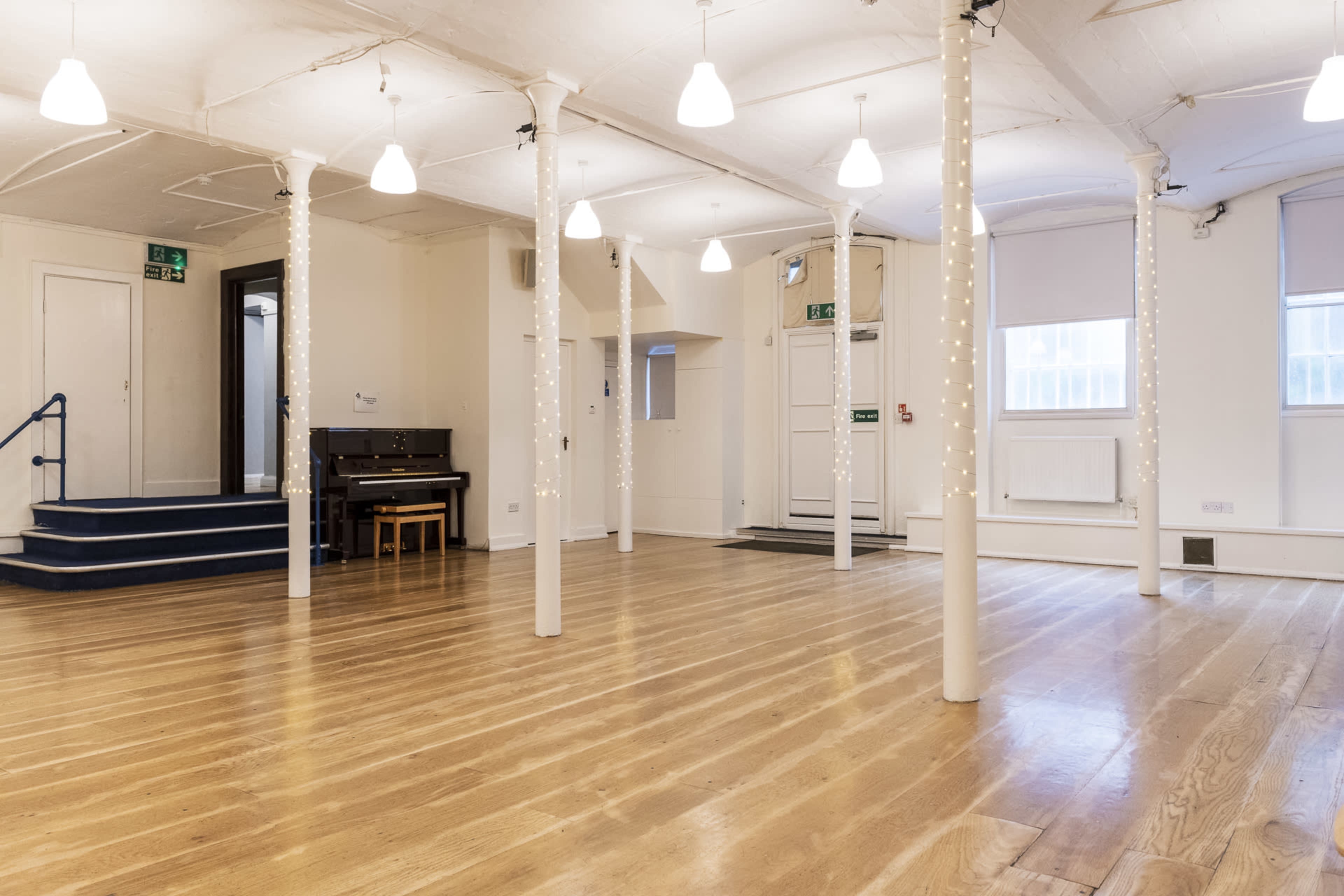 A spacious, well-lit room with wooden flooring, white pillars decorated with string lights, and a small piano against one wall.