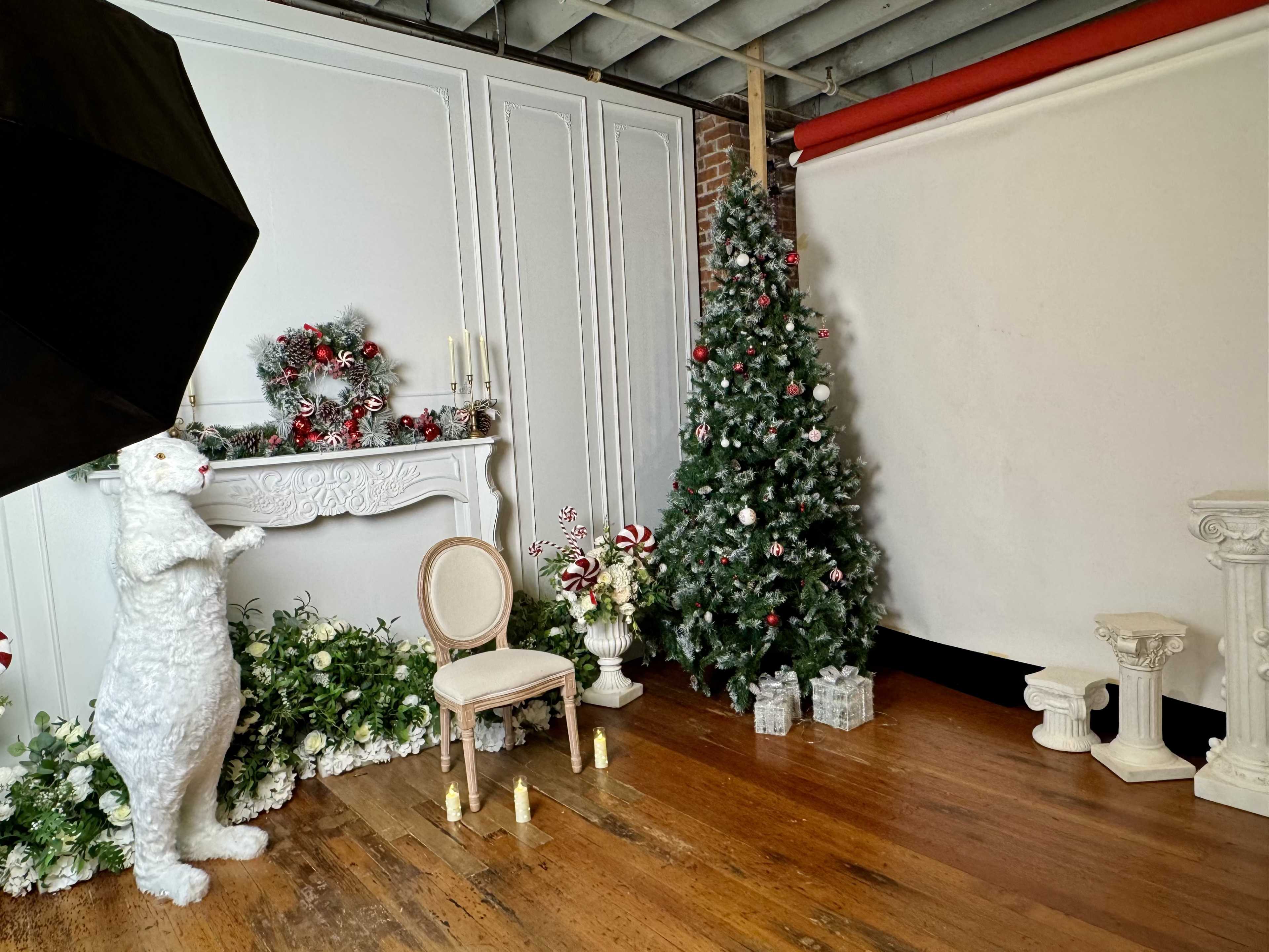 The scene features a decorated Christmas tree, a white fireplace adorned with festive decor, a chair, and a large white rabbit sculpture in a studio setting.