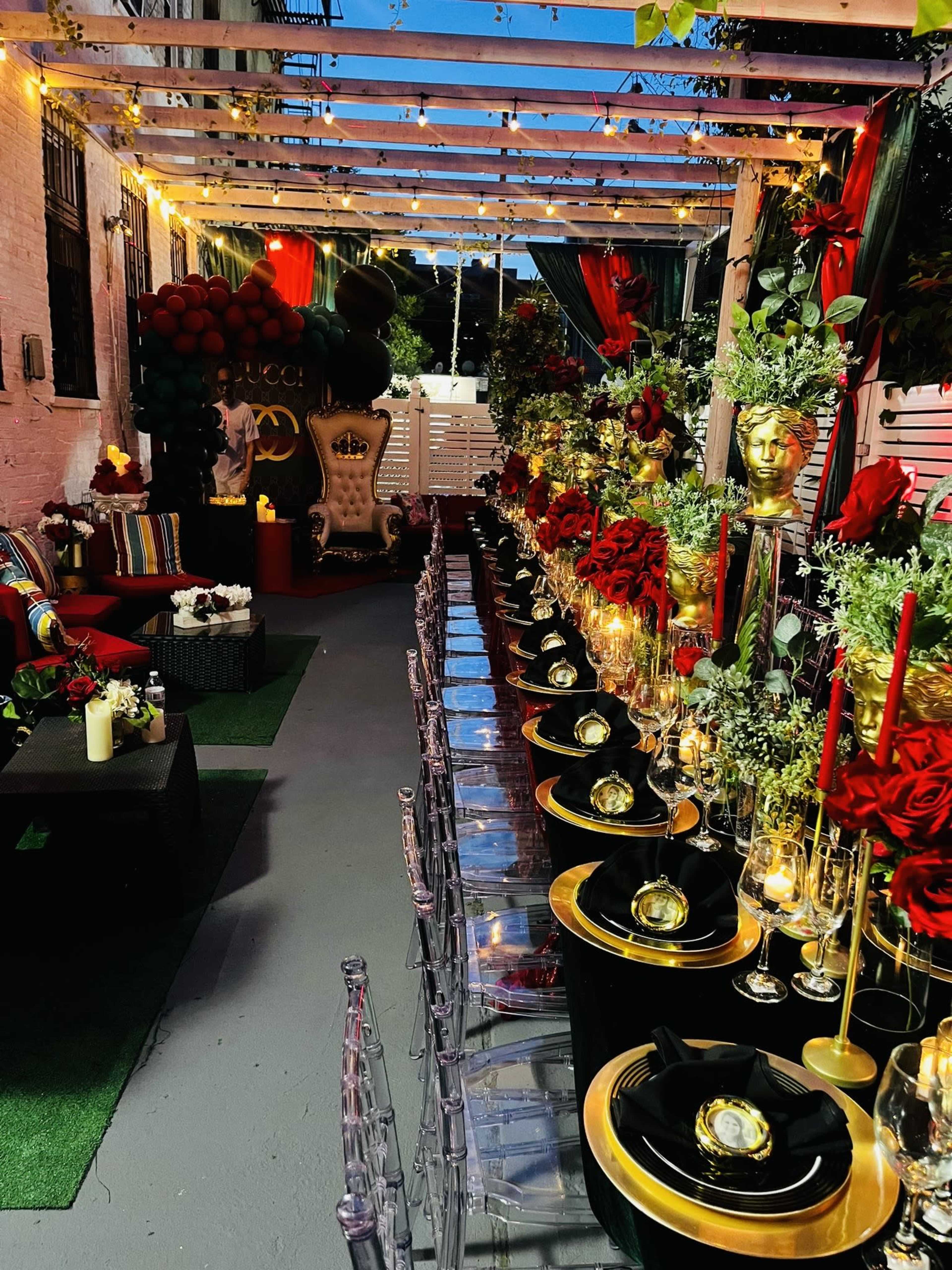 The image depicts a lavishly decorated outdoor dining area with a long table set for a formal event, featuring gold and black tableware, red roses, and elegant lighting.