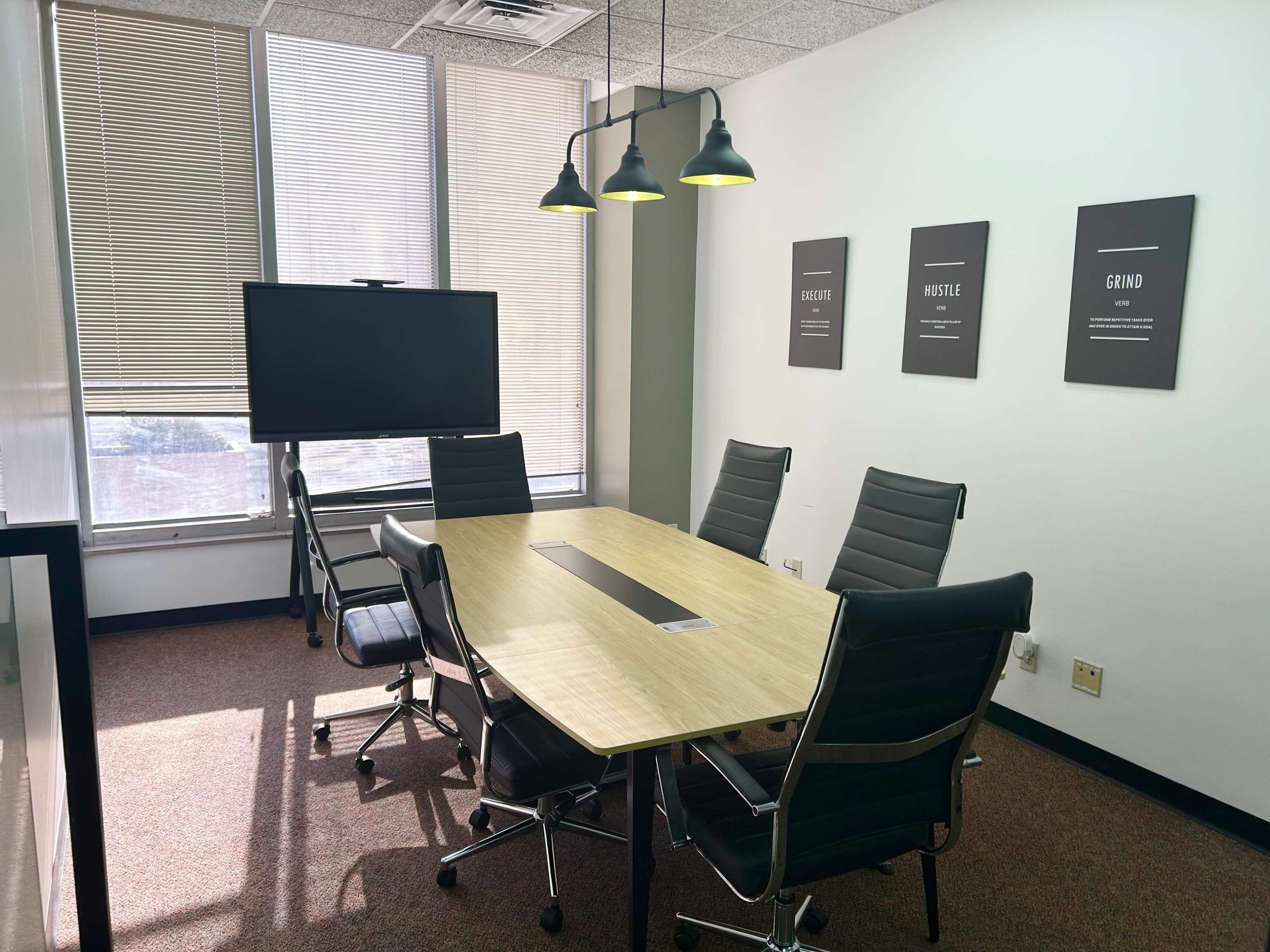 The image shows a conference room with a wooden table surrounded by six black office chairs, a television mounted on the wall, and framed posters hanging above the table.