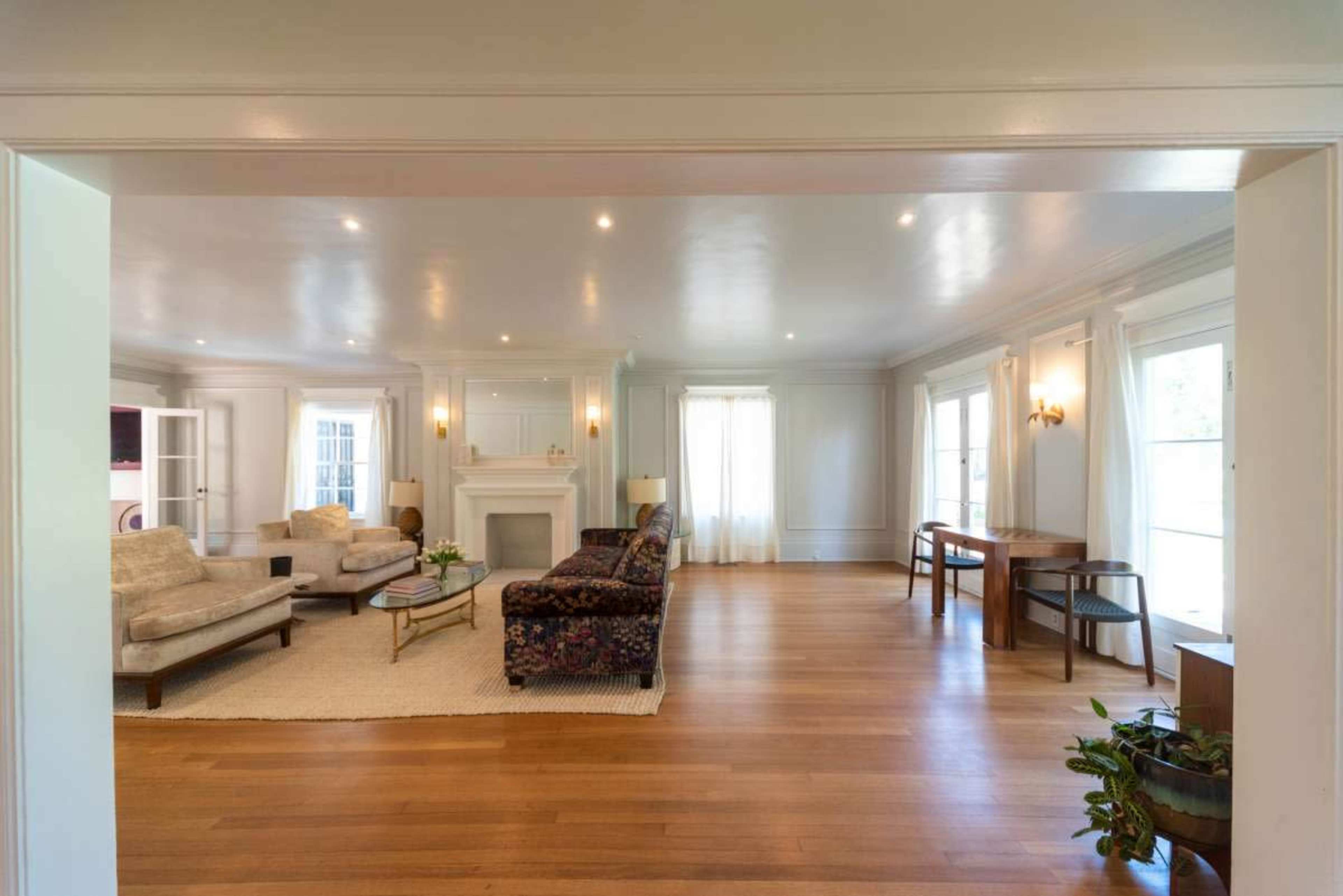 The image shows a spacious living room with two seating areas, a fireplace, and large windows allowing natural light to fill the space.