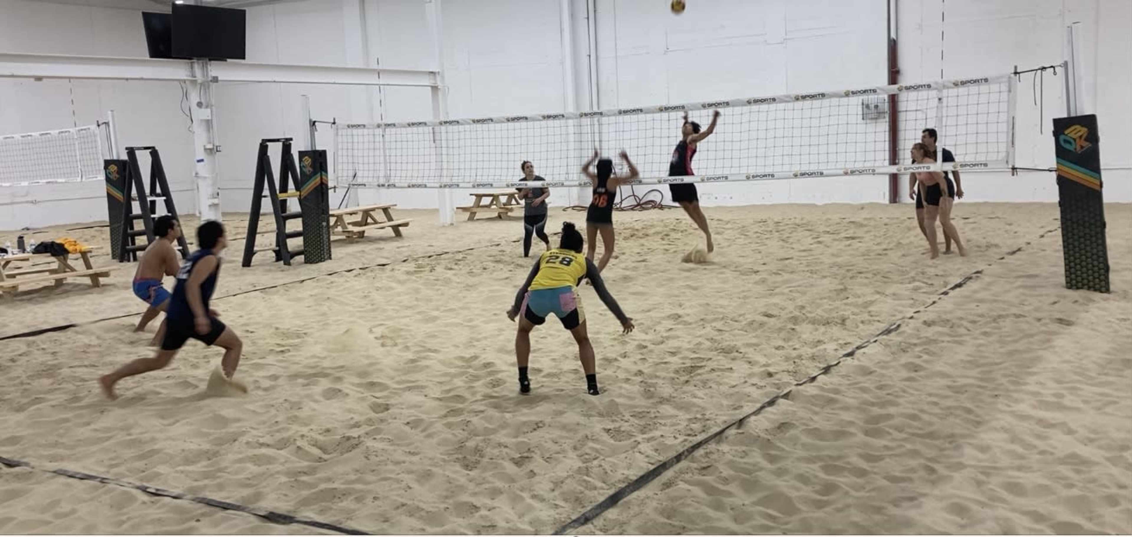 A group of six people plays beach volleyball indoors on a sand court.