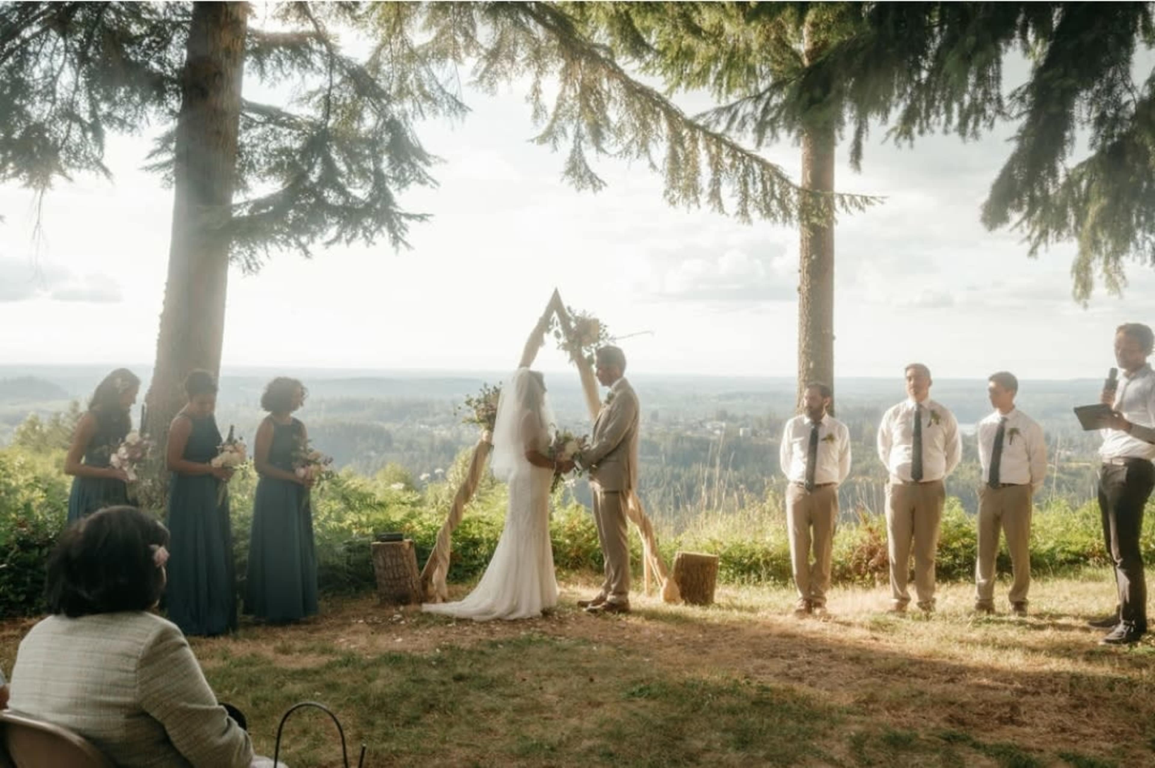 A couple stands under an arch made of wood and greenery, exchanging vows in front of guests at an outdoor wedding surrounded by trees and a scenic view.