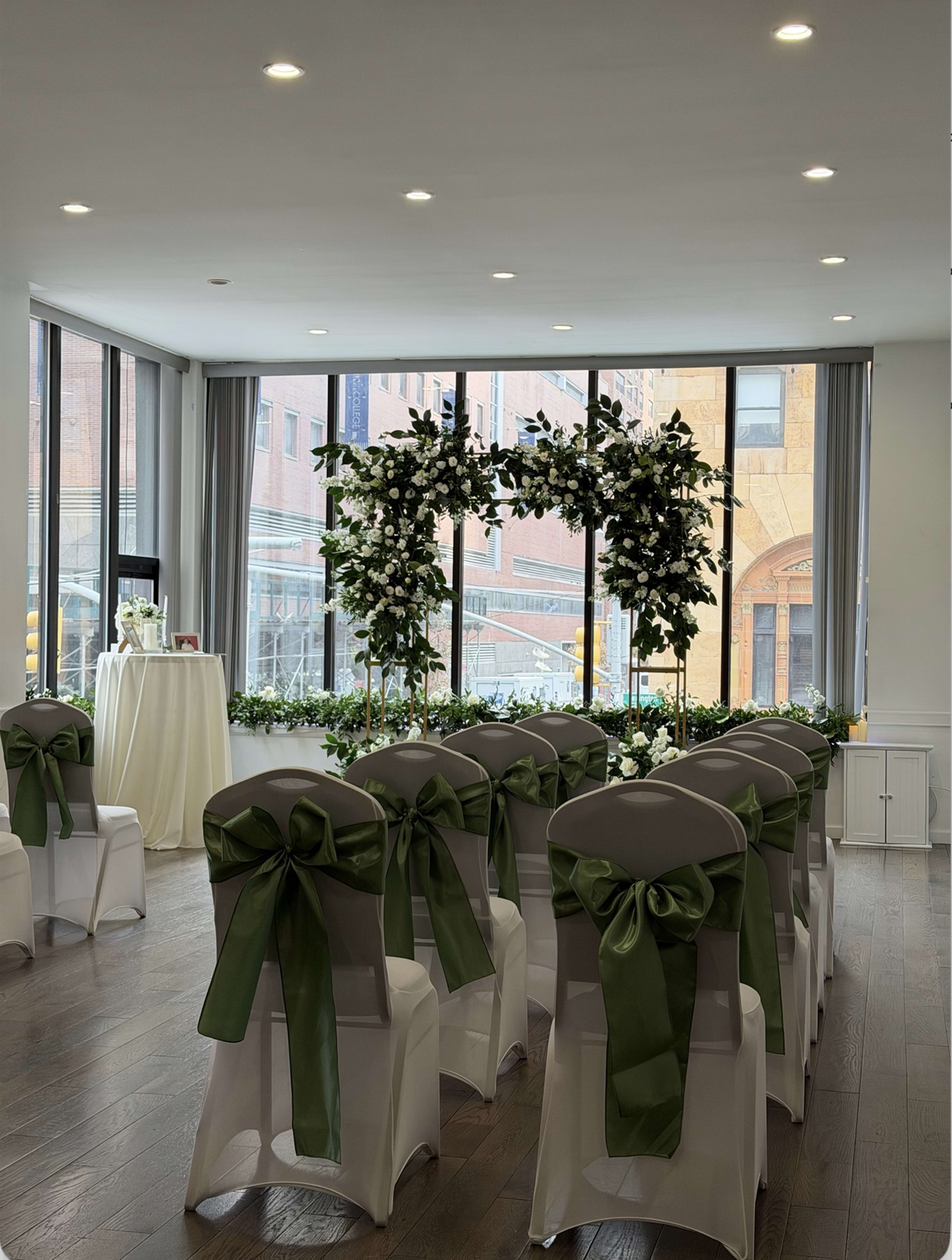 The image shows a wedding ceremony setup with white chairs adorned with green sashes, facing a floral arch under large windows.