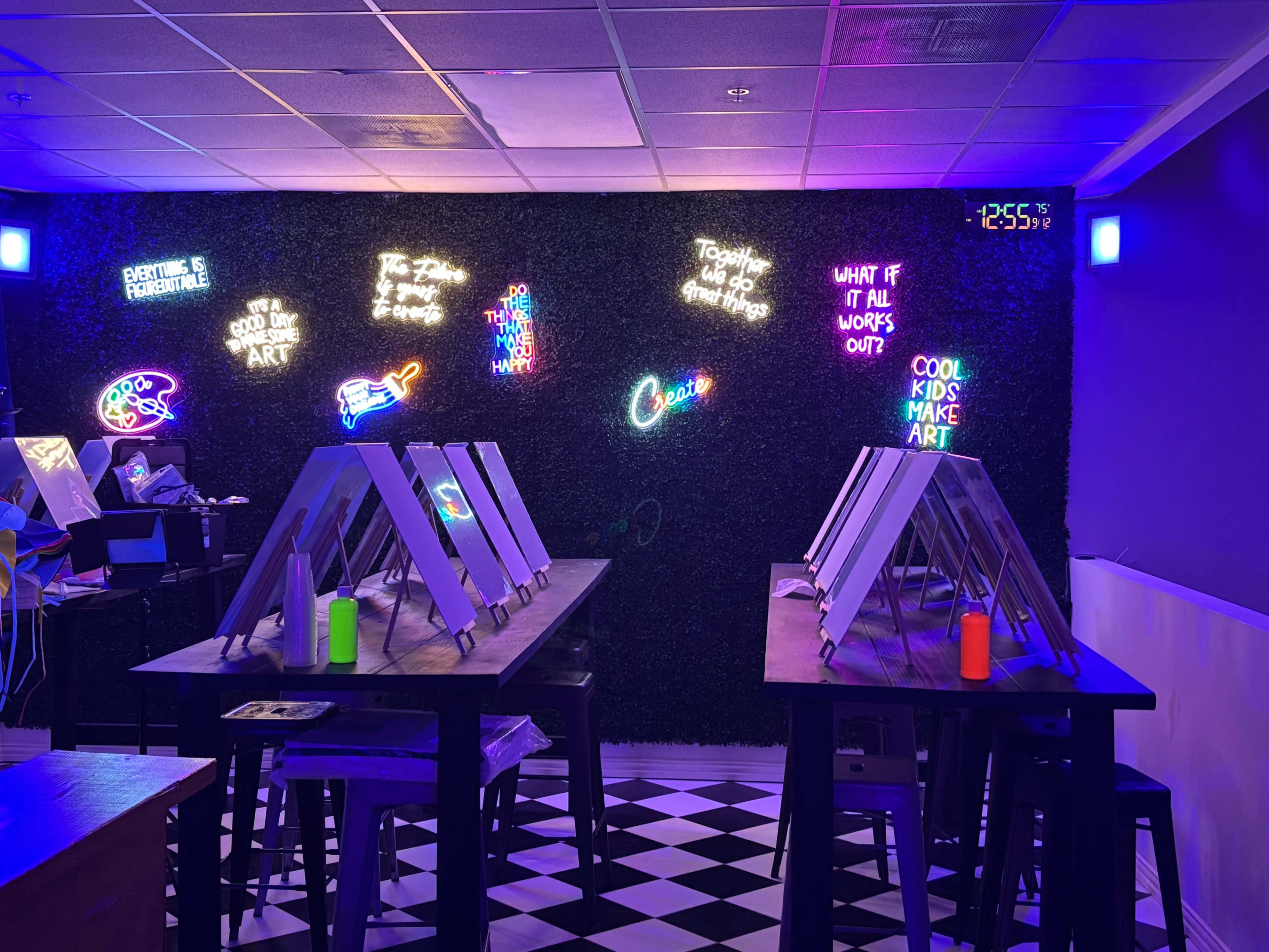 The image shows an art studio with several easels set up on tables, illuminated by colorful neon signs on a dark green wall.